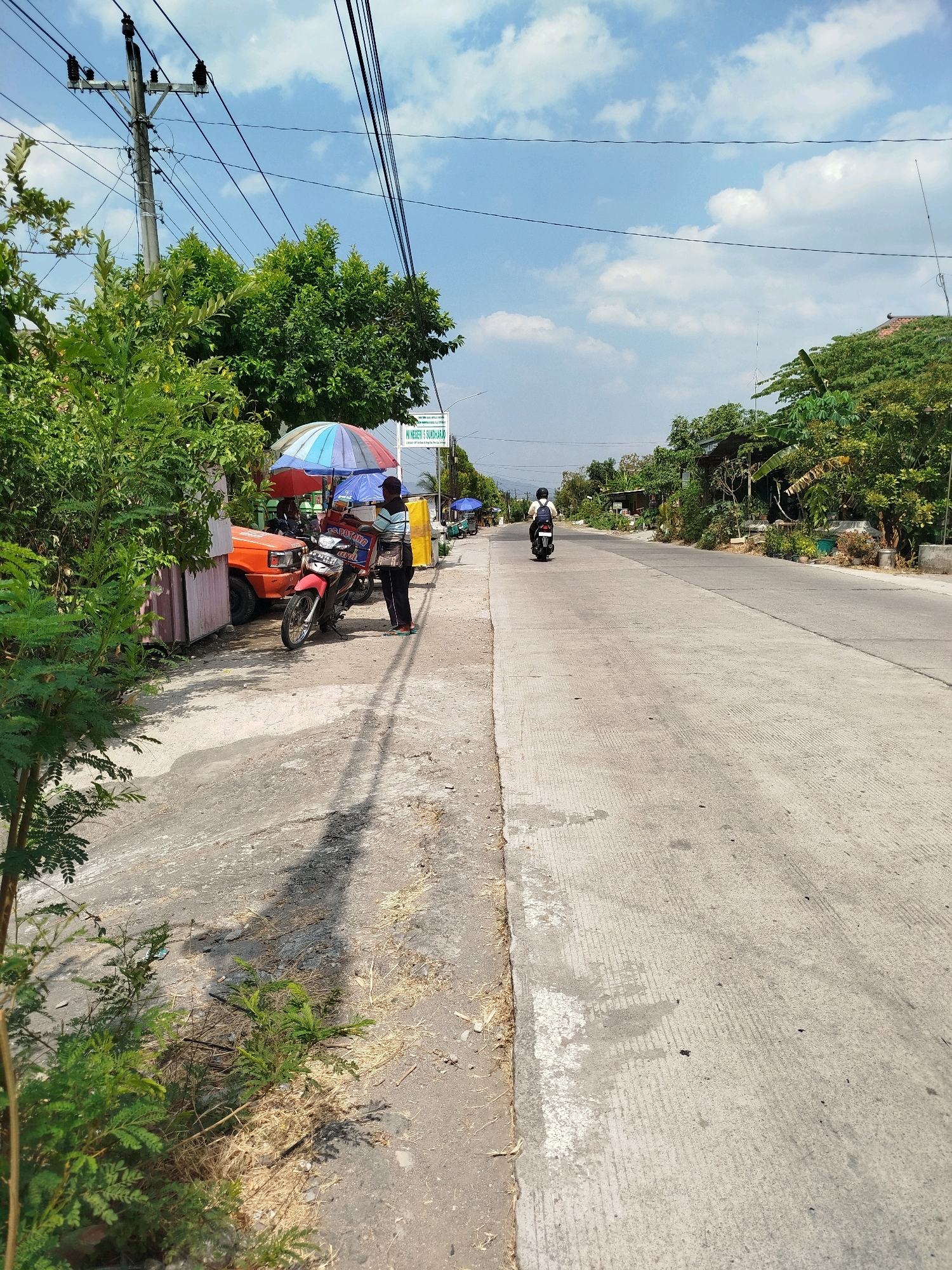 The image depicts a street scene in Grogol, Indonesia, on a sunny day with scattered clouds. A paved road runs through the frame, bordered by a sidewalk and lush green trees.
On the left side of the road, a small orange truck is parked next to a stall covered by a colorful umbrella. A person stands beside a red motorcycle, which has a brown cooler attached to its rear. Another person is seated on the motorcycle, and a third person is visible further down the sidewalk, near another stall with a blue umbrella.
Further down the road, two individuals are riding motorcycles. One motorcycle, ridden by a person wearing a helmet and backpack, is closer to the viewer and heading away from the camera. Another motorcycle is further in the distance. A sign is visible on the right side of the road, indicating a school: "SM NEGERI 5 SUNDARI". Power lines crisscross the sky, supported by a utility pole on the left. The ground is a mixture of concrete and dirt, with dry grass and foliage present. The overall impression is a bustling but calm street environment in a tropical setting.