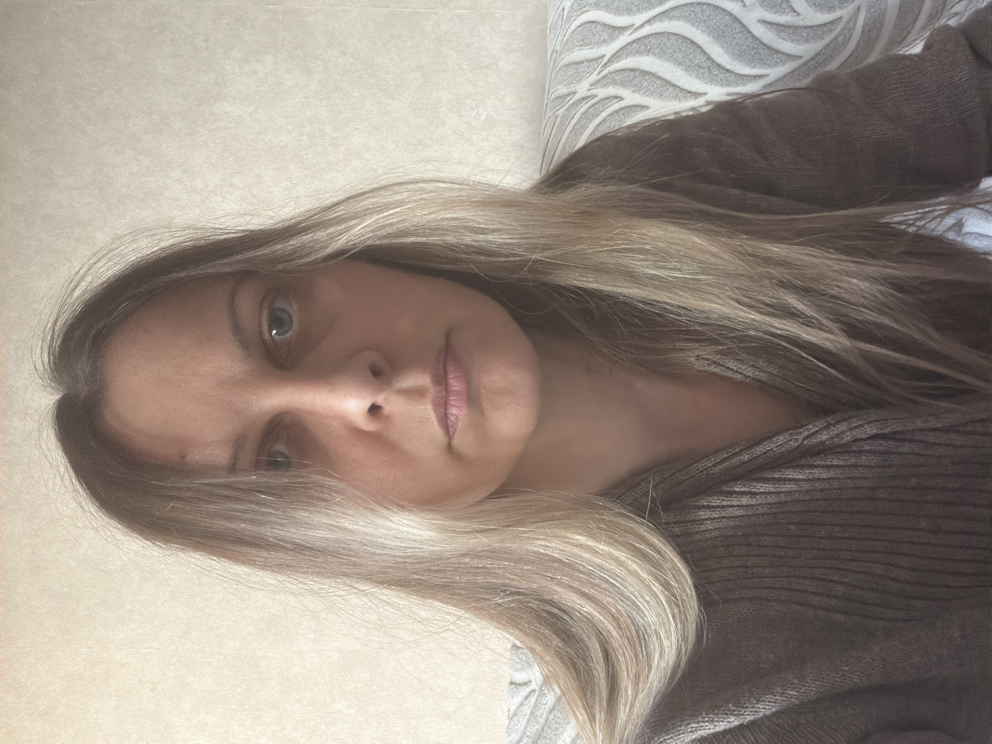 This rotated portrait features a woman with long, straight, ash-blonde hair and visible darker roots. She looks directly at the camera with light-colored, possibly blue or green, eyes. Her expression is neutral to slightly serious, and her lips are naturally pink. She appears to be wearing a dark, possibly brown or grey, ribbed or knitted sweater.
The setting is indoors, with a plain, light-colored wall visible on one side and a patterned surface behind her head and shoulder. This patterned surface features a repeating, abstract wavy or leaf-like design in light grey or white against a similar light background, possibly a wallpaper or headboard. The lighting is soft and even, suggesting an indoor environment, likely during the day or under ambient artificial light. No specific time of day or weather conditions are discernible.
The woman is posing for the photograph. The image provides no specific location cues to confirm the external context of Izhevsk, Russia, beyond the subject's appearance in an indoor domestic setting. There is no visible text in the image.