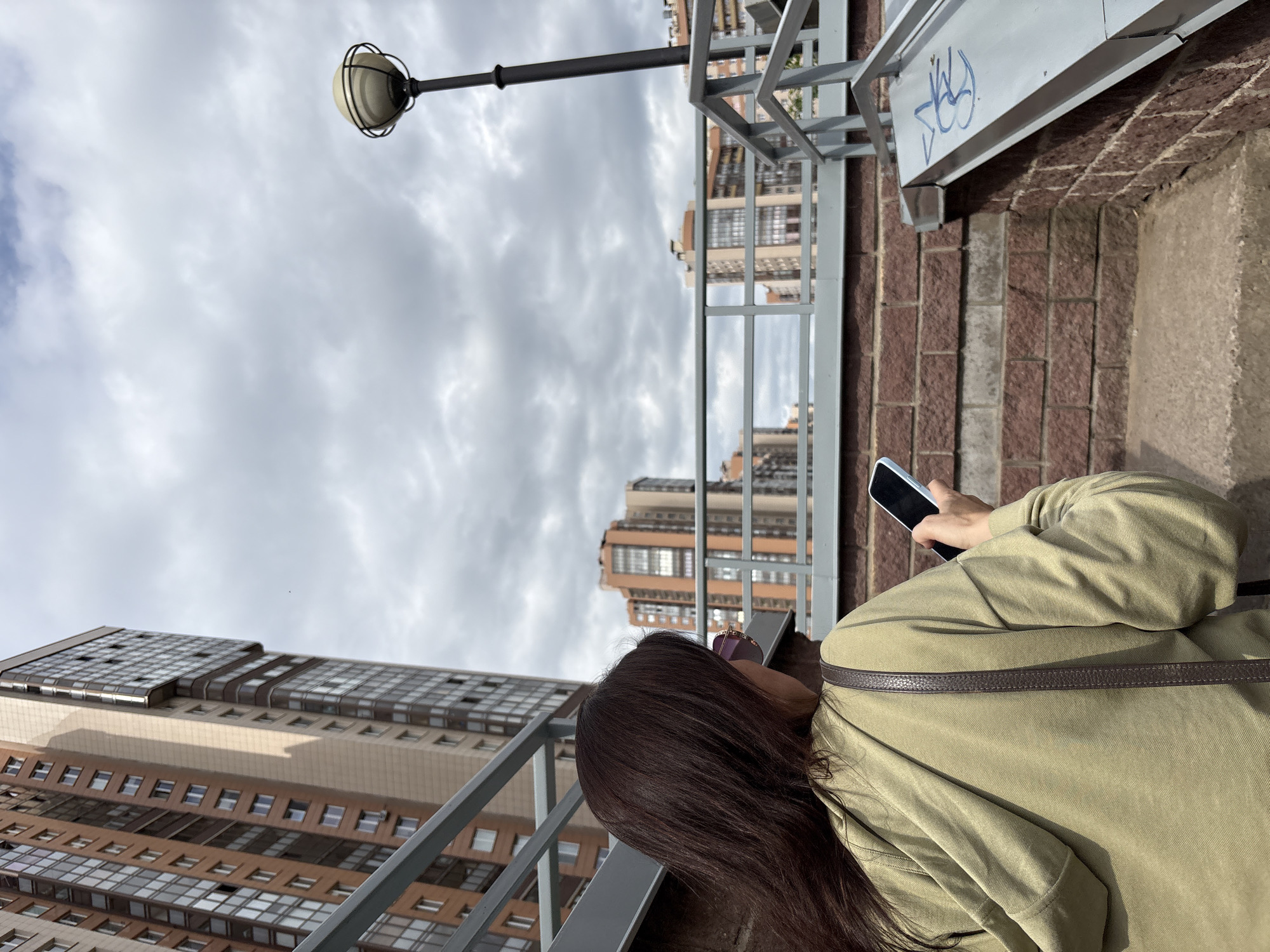 The image, viewed in its correct orientation, depicts an urban landscape from a slightly elevated vantage point, likely a bridge, overpass, or high balcony, in Saint Petersburg, Russia. A person with long, dark brown hair is seen from behind and to the right, wearing a light olive green long-sleeved jacket and a dark brown strap across their shoulder. They are holding a white or light-colored smartphone in their right hand, seemingly engrossed in its screen.
Grey metal railings stretch across the foreground and mid-ground, separating the viewer from the distant city buildings. On a grey metal beam or structure to the right, a distinctive piece of blue graffiti, possibly "VLKV" or similar stylized lettering, is visible. A black lamp post with a spherical, caged light fixture rises into the sky from the upper left.
The background showcases several modern multi-story buildings, characteristic of a contemporary city. These structures feature varying shades of brown, beige, and grey with numerous windows, reflecting the predominantly overcast sky. One prominent building on the left has a unique stepped or multi-tiered roof design. The sky is filled with soft grey clouds, indicating a cloudy daytime. The scene captures a quiet moment of an individual engaging with technology amidst the urban environment.