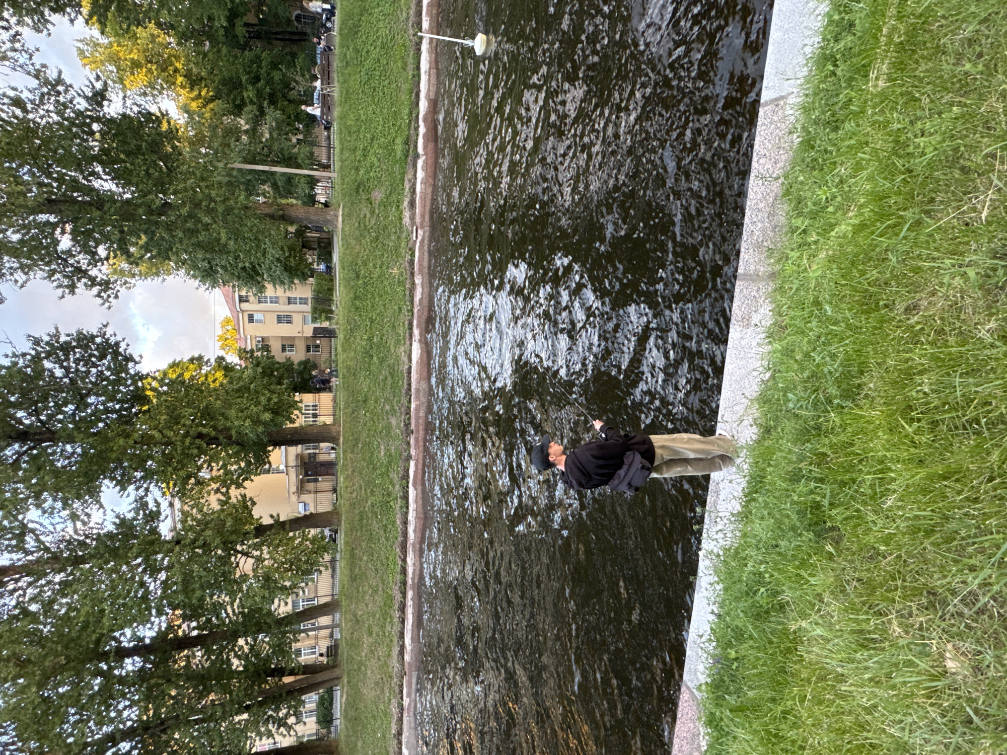 A man wearing a black hoodie, khaki pants, and a black baseball cap is fishing on the edge of a canal in Saint Petersburg, Russia. He has a dark-colored bag slung across his chest. The canal is dark and rippling, with reflections of trees on its surface. To the left of the canal, there is a grassy embankment, and further back, there are tall trees, a sidewalk, a fence, and residential buildings. The sky is visible between the trees, showing some clouds. On the canal, a white buoy with a yellow base is visible in the distance.