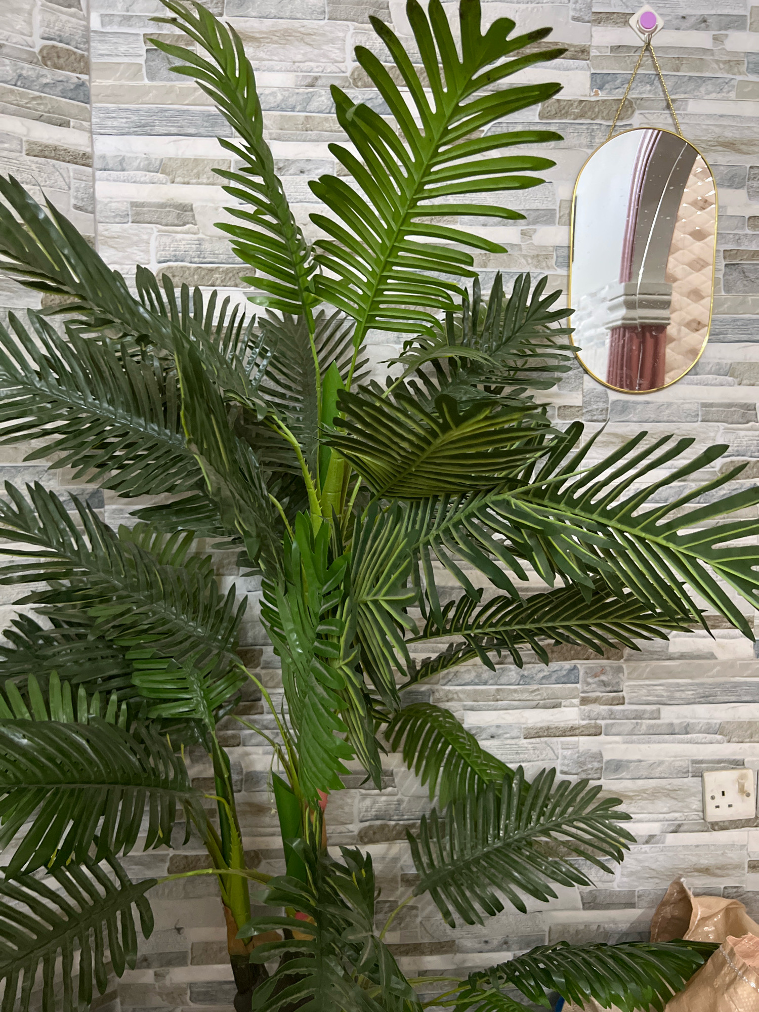 A tall, artificial palm plant with dark green fronds is positioned in front of a grey and white brick-patterned wall. To the right of the plant, a gold-rimmed, oval mirror hangs on the wall, reflecting an interior scene with a curved architectural element and columns. Below the mirror, a white electrical outlet with two sockets is visible on the wall. In the bottom right corner of the image, part of a light brown bag or packaging is visible. The scene appears to be indoors, potentially in a domestic or commercial setting in Dar es Salaam, Tanzania.