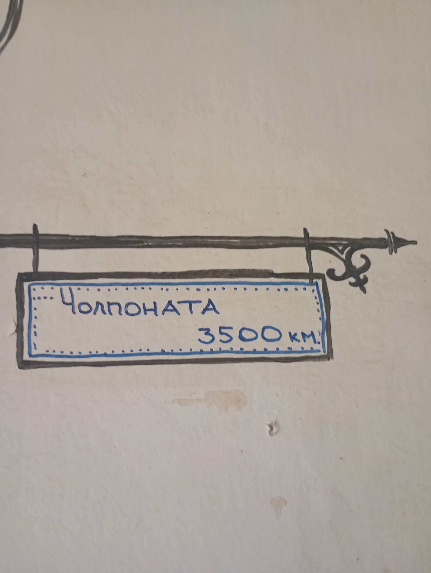The image shows a hand-drawn sign on a textured white wall. The sign is rectangular with a dark gray border and a blue dotted inner border. The text on the sign is written in blue marker. The top line reads "ЧолПOНАТА" and the bottom line reads "3500 км.". The sign is suspended from a dark gray horizontal bar, which is attached to a decorative dark gray wrought-iron bracket. The background is a plain white wall with some subtle discolorations. There are no people or other objects visible in the image. The image does not provide any information about a specific location in the city of Moscow, Russia.