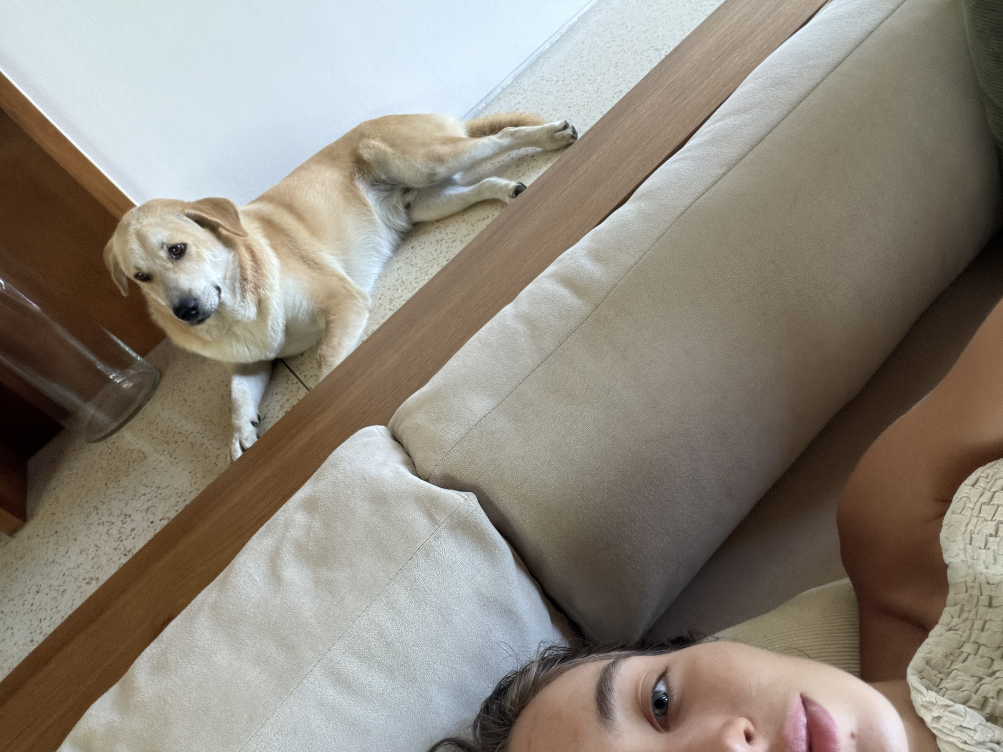 The image shows a woman lying on a light-colored couch, with the camera angled upwards towards her face. The woman has fair skin, dark hair, and is partially visible from the neck up. A light-tan colored dog is lying on the floor beside the couch, looking towards the camera. The floor is a light-colored, speckled material. A wooden edge runs along the side of the couch, separating it from the floor. A glass bottle is visible near the lower left corner, next to a wooden structure. The location is possibly in Mas, Indonesia, but this cannot be determined with certainty from the visual elements in the image.