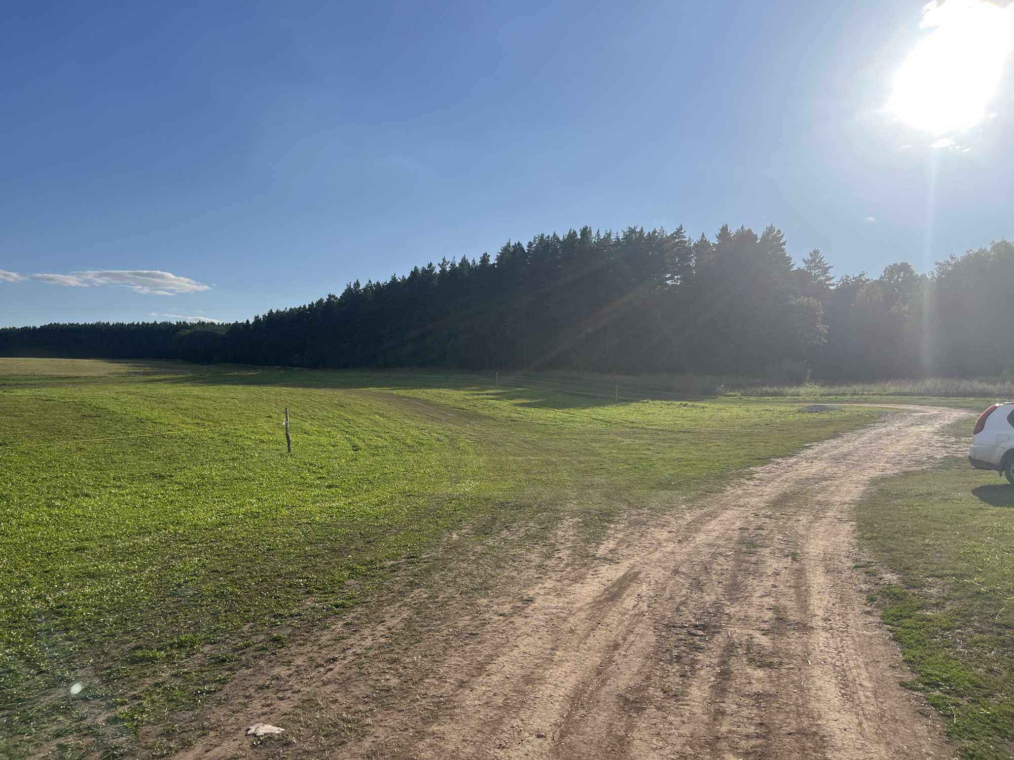 This is a landscape photograph taken on a bright, sunny day in Srednie Pinyachi, Russia. The image depicts a dirt road curving through a grassy field bordered by a dense forest. The sky is a clear blue with a few wispy clouds, and the sun is shining brightly, casting lens flares and light rays across the scene. In the foreground, the dirt road is well-worn with visible tire tracks. To the left, the field is covered in lush green grass, with a single wooden post sticking out of the ground. The forest, a mix of evergreen trees, forms a dark line along the horizon, with the sun's rays partially obscured by the trees on the right side of the frame. A white SUV is partially visible on the right edge of the image, parked on the grass next to the dirt road. The overall impression is one of a natural, rural setting on a warm, clear day.