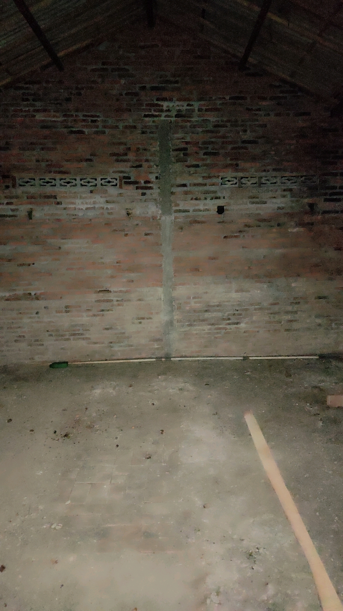 The image depicts the interior of a partially constructed or unfinished building in Tanjungan, Indonesia. The walls are made of red brick, with a central vertical seam that appears to be concrete. There are decorative patterns of alternating brick and gaps along the upper portion of the walls. The floor is concrete, with visible tiling underneath in some areas. A corrugated metal roof is visible at the top of the frame, supported by wooden beams. There is a long, light-colored wooden plank lying diagonally across the floor in the foreground. The scene is dimly lit and appears to be in a state of disuse or abandonment. There are no people or specific actions taking place.