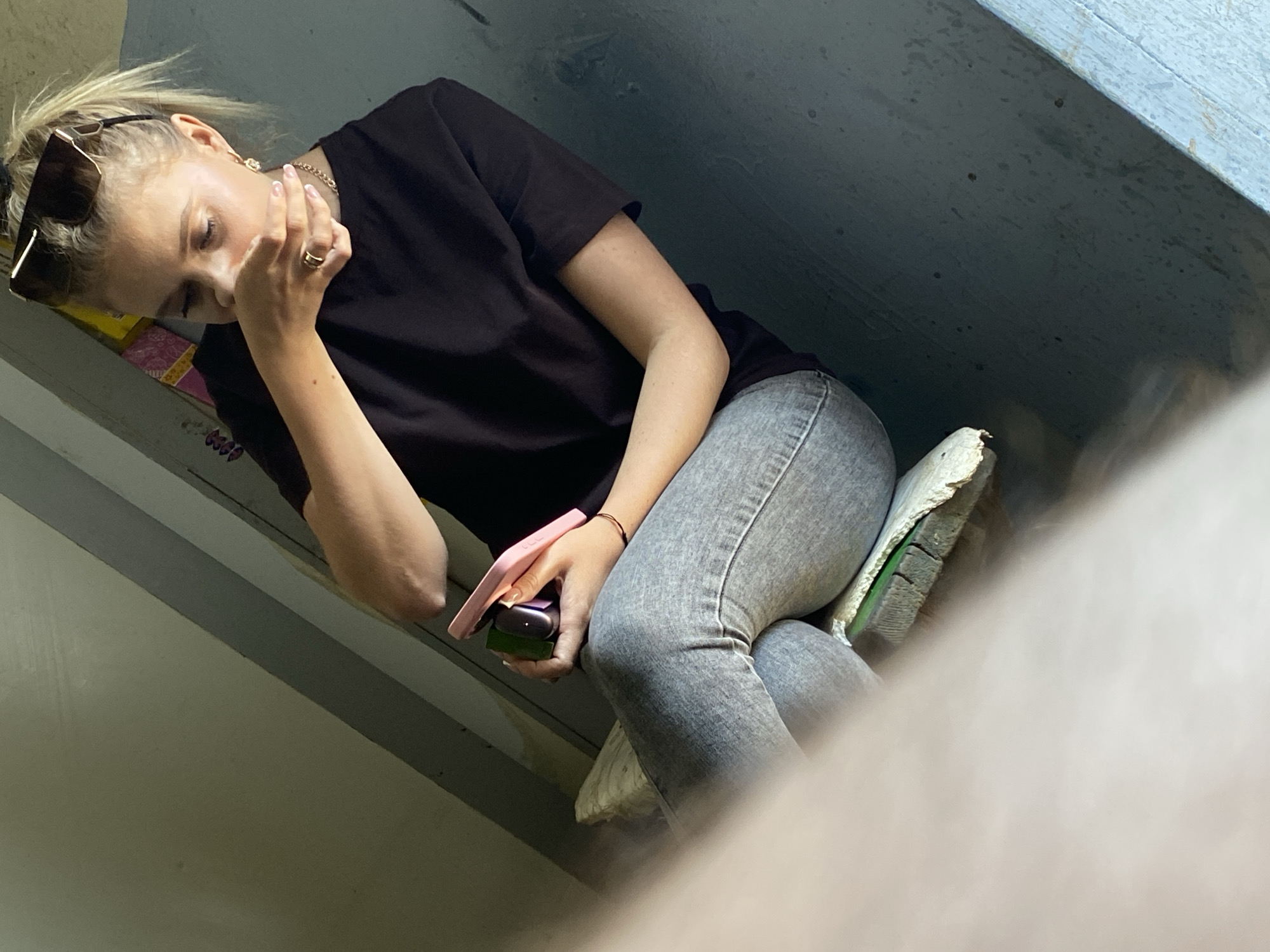 A young, blonde woman with sunglasses on her head sits on a bench. She is wearing a black t-shirt and light-wash jeans. She is holding a pink phone and a green electronic device in her hands. Her right hand is covering her mouth. She is indoors, and the background appears to be a dimly lit, possibly industrial or utility space with gray walls and some yellow and pink patterned fabric visible. The lighting suggests it may be daytime.
