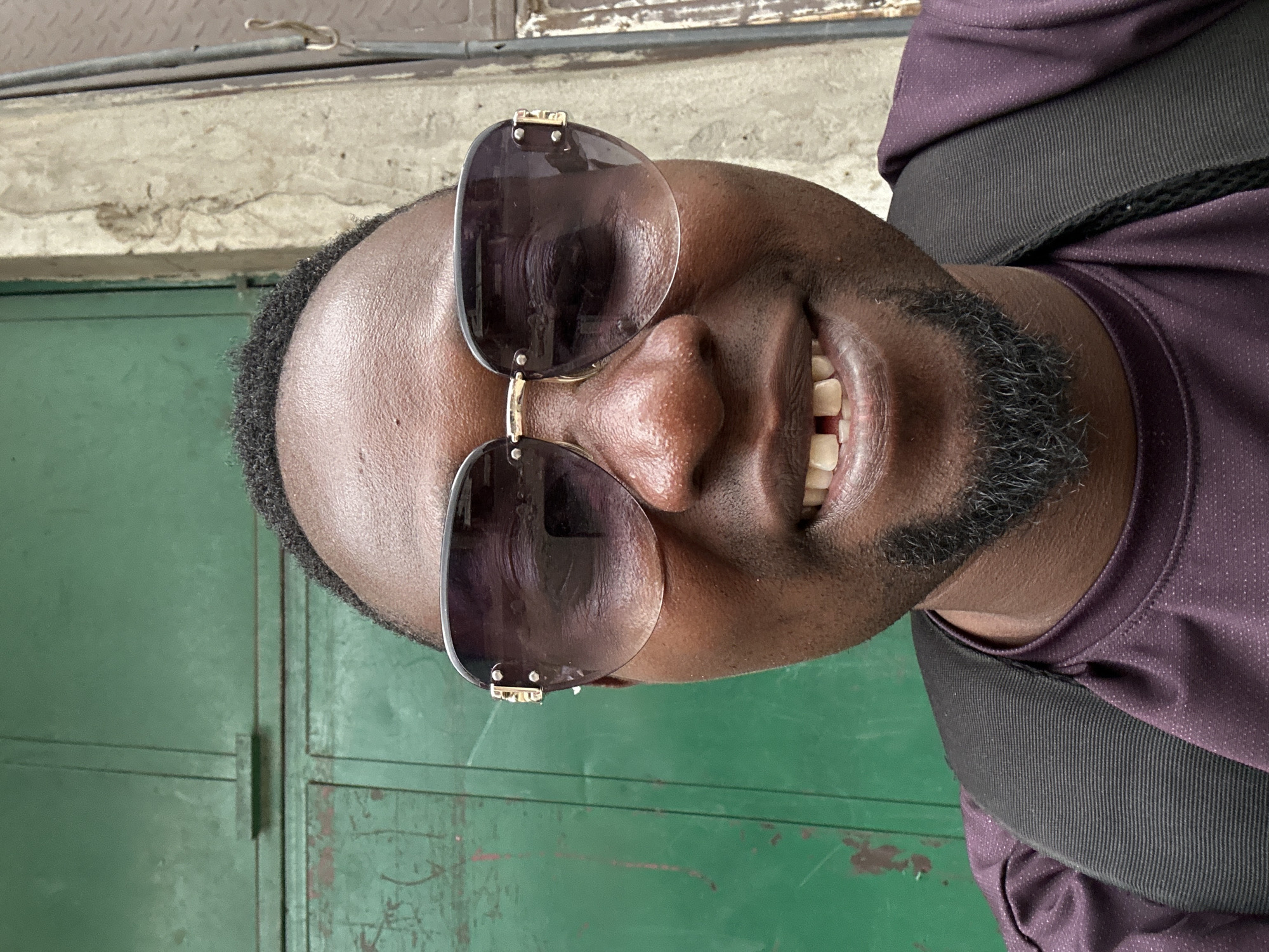 A vibrant, close-up portrait depicts a Black man smiling broadly, facing slightly to the viewer's right. He wears large, dark-lensed sunglasses with gold-colored accents on the frames, obscuring his eyes. His wide smile reveals healthy teeth and gums, conveying a cheerful and positive emotion. He has short, dark, curly hair and a neatly trimmed goatee-style beard.
He is dressed in a dark purple short-sleeved shirt made of a textured, possibly athletic, fabric. A black strap, likely from a backpack or bag, crosses over his left shoulder.
The background suggests an outdoor or urban setting. Directly behind him is a green metal surface, possibly a door or wall, showing some signs of wear and age. Above this, a lighter-toned concrete or stone ledge is visible, with a thin metal bar or pipe running horizontally across the upper portion of the frame. The bright lighting indicates it is daytime. There is no visible text in the image.