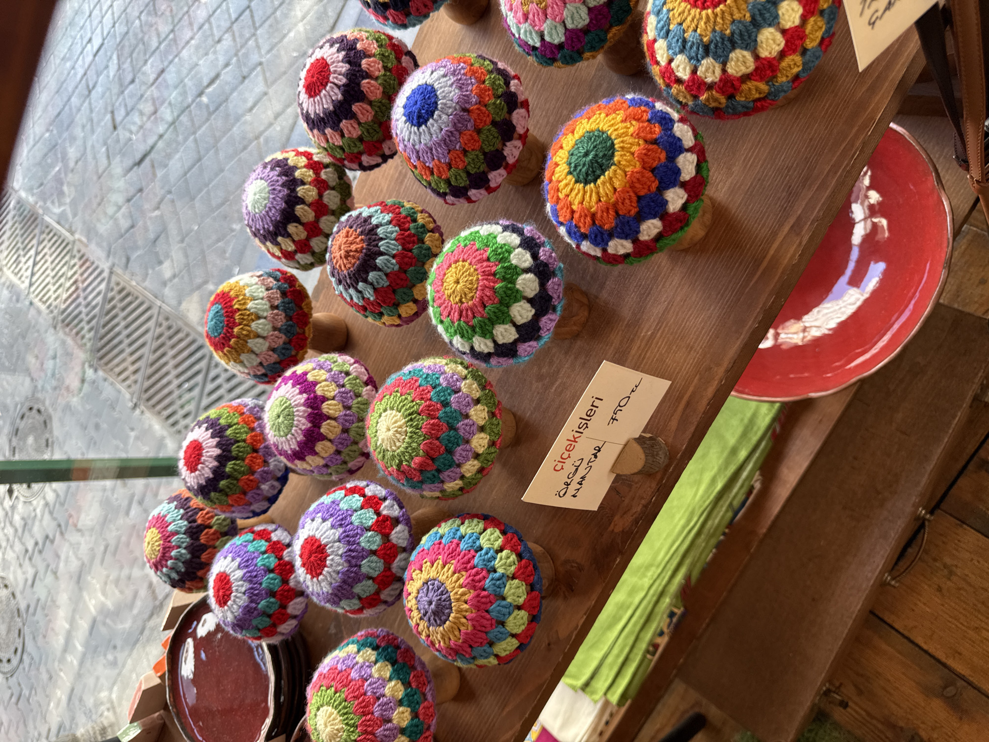 In Kadıköy, Turkey, a collection of hand-crocheted mushroom-shaped ornaments is displayed on a wooden shelf. The ornaments feature vibrant, multicolored patterns created with yarn in shades of red, purple, yellow, green, blue, pink, and orange. Each ornament is mounted on a small wooden base. To the left, a stack of reddish-brown ceramic plates is visible, along with a folded green fabric item. A small white sign with handwritten text in Turkish, likely indicating the price and type of item, is propped up among the ornaments. The background suggests an outdoor setting with a tiled floor and what appears to be a window with reflections of the sky and the street. The overall scene captures a display of artisanal crafts in a marketplace or shop.