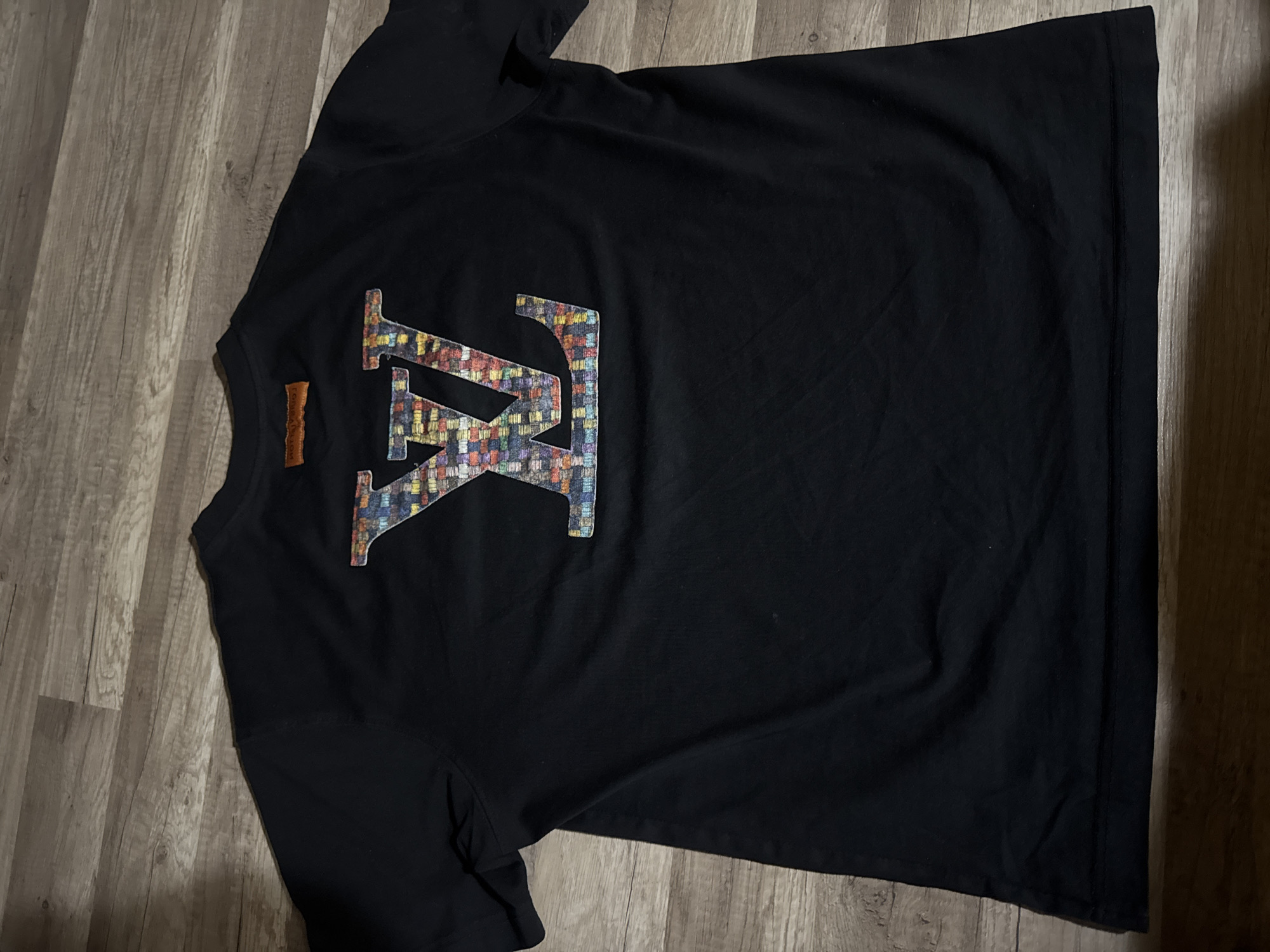 A black T-shirt is lying on a wood-patterned floor. The T-shirt has a large design on the upper back consisting of two overlapping letters. The letters have a multicolored, woven or embroidered appearance. There's also a small, orange rectangular tag on the back near the neck. The T-shirt is wrinkled. The setting is indoors.