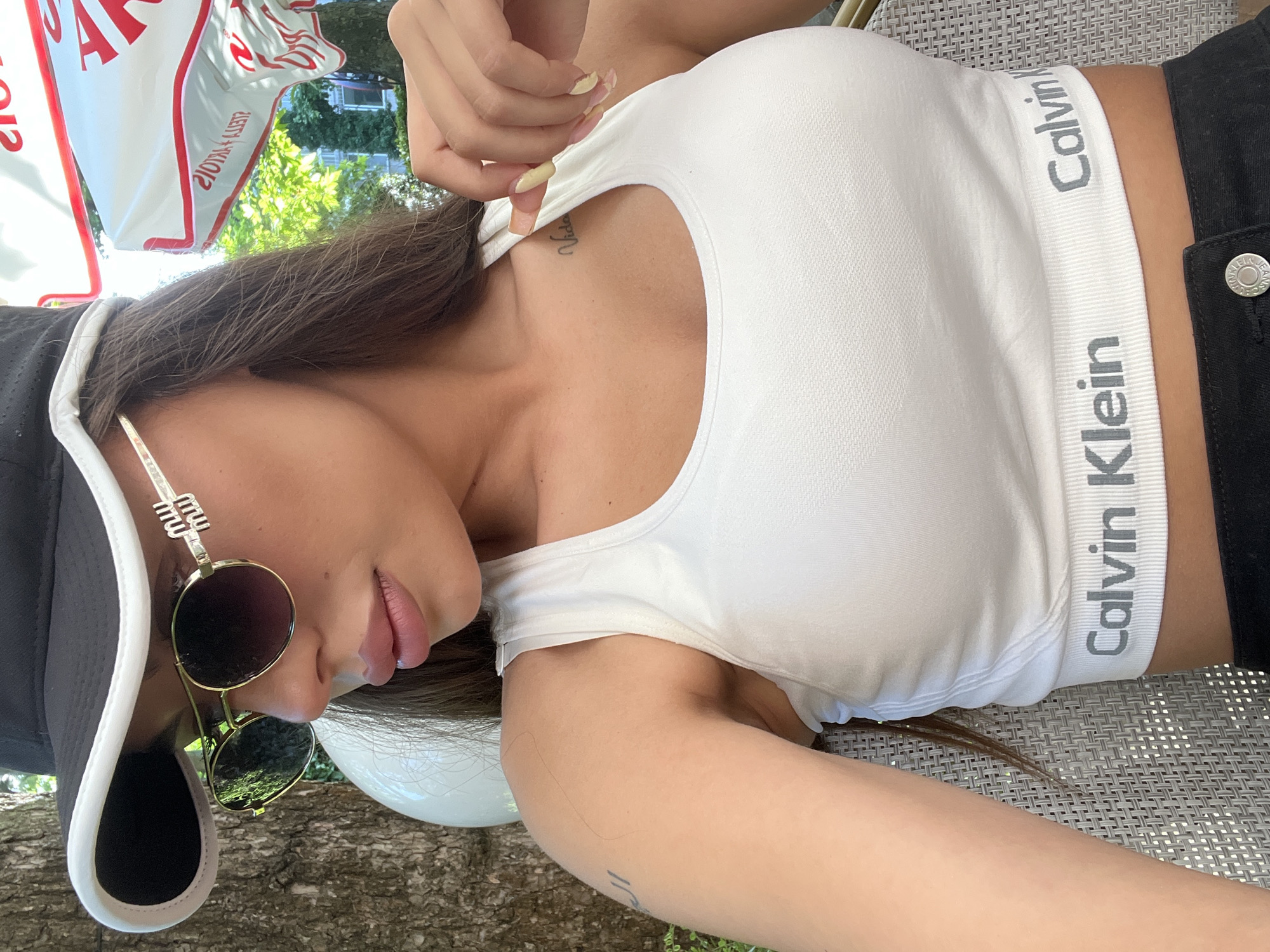 A woman is depicted sitting outdoors at a cafe or restaurant in Burgas, Bulgaria. She is seen from the chest up, positioned slightly to her left. She wears a black baseball cap with a white brim, gold-rimmed aviator-style sunglasses with dark lenses and a distinctive "MW" emblem on the temples, and a white Calvin Klein sports bra/crop top featuring "Calvin Klein" printed in grey on the elastic band. Her long, dark brown hair frames her face and falls over her shoulders. Her lips are full with light pink lipstick, and her long nails are painted light yellow/cream. Tattoos are visible on her upper right chest, reading "Vida," and abstract script on her left bicep. Her right hand is lightly touching the strap of her top. She is seated in a light grey woven rattan-style chair. The background to her left features a tree with textured bark and green foliage. To her right, a large white outdoor umbrella displays red Cyrillic text, including "СТОЛОВЕ" (STOLOVE), indicative of a Bulgarian establishment. Further back, additional green trees and distant buildings are visible under bright daylight.