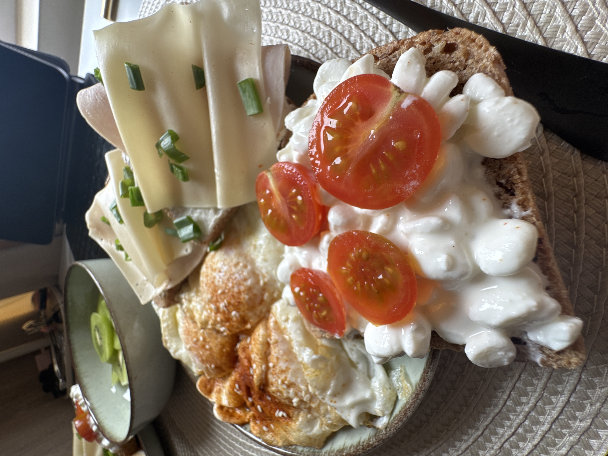 A close-up, angled, high-angle shot of a plate of food. The food consists of a slice of bread with cottage cheese and sliced tomatoes, topped with egg and cheese slices with chives. To the left is a bowl containing sliced kiwi fruit. The plate sits on a textured mat. A knife is on the top right of the plate. The location is Sofia, Bulgaria.