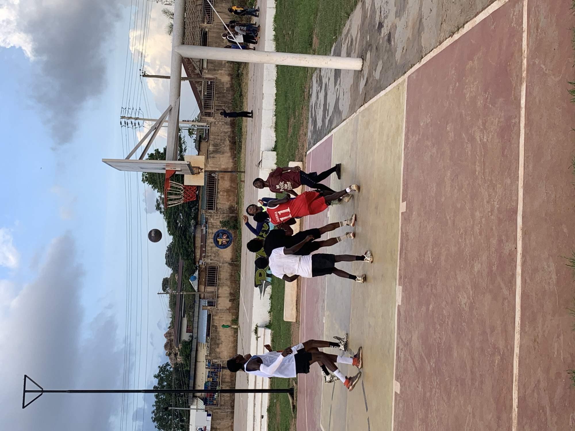 A group of young men are playing basketball on an outdoor court in Gura Top, Nigeria. The court has a reddish-brown and yellow surface with white lines. A basketball hoop with a red net is visible. The sky is overcast with some clouds. In the background, there are buildings, trees, and power lines. Several players are in motion, some reaching for the ball. One player in a red jersey with the number 11 is prominent. Another player in a white t-shirt and black shorts is also visible. The scene suggests a casual game or practice session. The lighting indicates it is likely late afternoon or early evening.