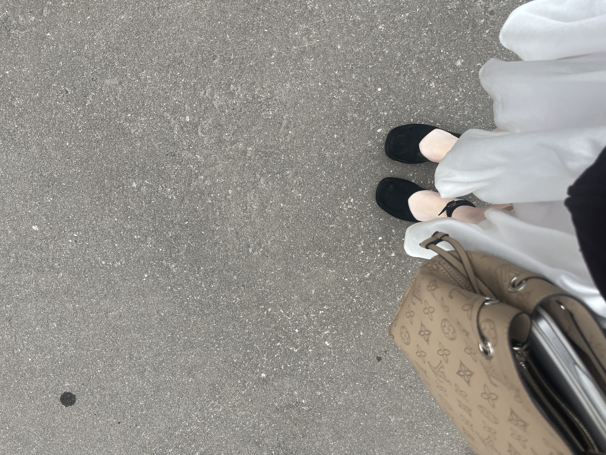 The image depicts a street scene from a first-person perspective, showing the lower part of a person's body. The person is wearing black flat shoes with a strap on the right foot and a flowing white dress. To the right of the person is a beige handbag with visible monogram patterns. The background consists of a gray asphalt surface. Based on the visual evidence available in the image, there is insufficient information to determine the specific location within Moscow, Russia.