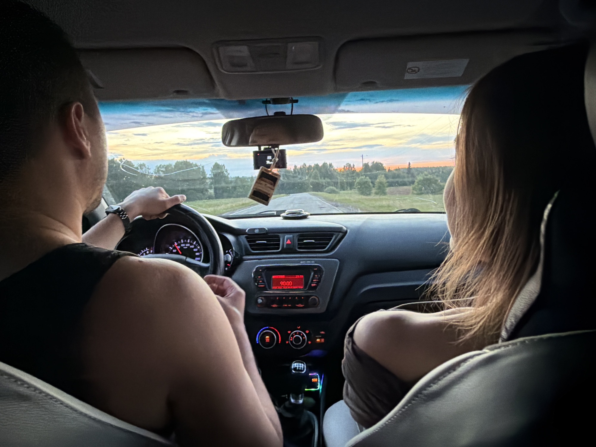 The image shows the interior of a car driving on a road near Mozhga, Russia. The driver is a man, viewed from the back, wearing a watch and a dark tank top. A woman with long, light brown hair is in the passenger seat. The car is driving towards the horizon, where the sky is a mix of blue, orange, and yellow, indicating either sunrise or sunset. The radio is tuned to 90.00. There is a rearview mirror with an air freshener hanging from it. Trees and fields are visible through the windshield.