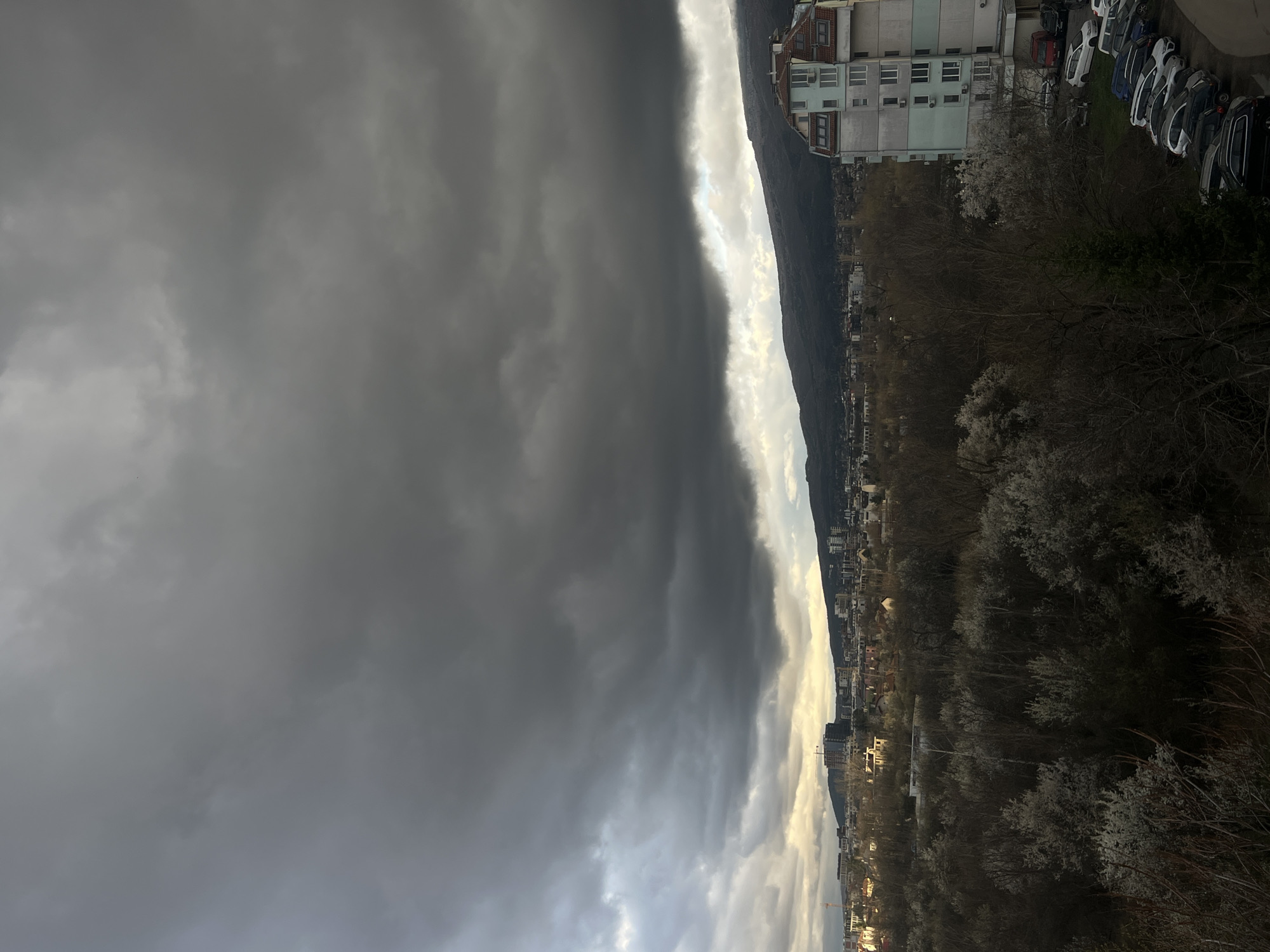The image captures a view of Sofia, Bulgaria, under a dramatic sky. A prominent mountain ridge forms the backdrop to the city. The sky is dominated by dark, ominous storm clouds on the left side of the frame. A lighter band of clouds or sky peeks between the storm clouds and the mountain ridge.
In the lower half of the image, a section of the city is visible. Buildings, trees (some with blossoms), and a line of parked cars are discernible. The light is dim, suggesting either late afternoon or an overcast day.