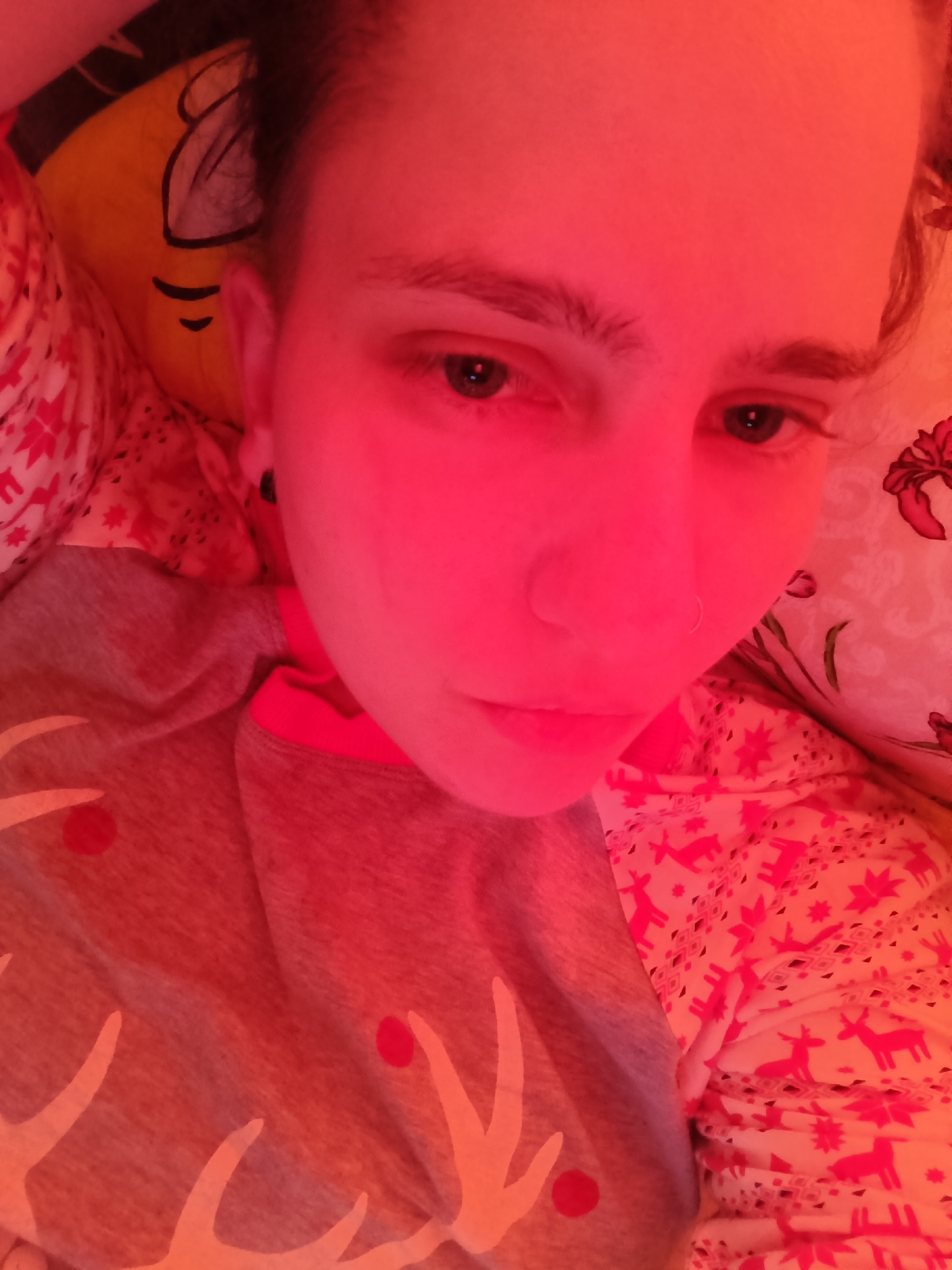 A person is seen lying down, likely in a bed, bathed in strong red-pink lighting that permeates the entire scene. Their head is resting on a yellow pillow with a black outlined facial feature, possibly a cartoon character. The person is wearing a grey long-sleeved top with white antler-like patterns and red circular designs, possibly representing Rudolph or a festive deer theme, with a pink trim visible around the collar.
The person's gaze is directed downwards. They have dark hair, pulled back, and visible eyebrows. A small silver hoop nose ring is present on their left nostril, and a dark earring is visible in their left ear. Their skin appears flushed red due to the intense lighting.
The bedding around the person includes a sheet or blanket patterned with numerous pink/red deer and snowflake-like motifs on a lighter background, further enhancing a potentially festive or cozy atmosphere. The overall impression is an intimate, quiet moment, heavily influenced by the distinct red illumination. There is no visible text in the image.