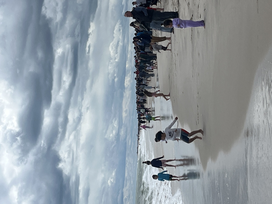 The image shows a beach scene in Wantagh, New York, at coordinates 40.592, -73.507. The sky is overcast with a dense layer of gray clouds. A large crowd of people is gathered on the beach, standing in a line along the shoreline. The people are facing the ocean, and most appear to be adults. A few children are playing in the shallow water and on the sand. Waves are breaking gently on the sandy shore. The beach is wide and covered with fine white sand. A young boy in a white t-shirt with the word "NIKE" is jumping in the shallows. Another young boy in a blue t-shirt and a young girl in a pink t-shirt are walking in the water. A young girl in a lavender outfit stands at the edge of the sand. There are a few adults behind the children on the beach. The beach is empty for the most part, with only a few people in the distance.