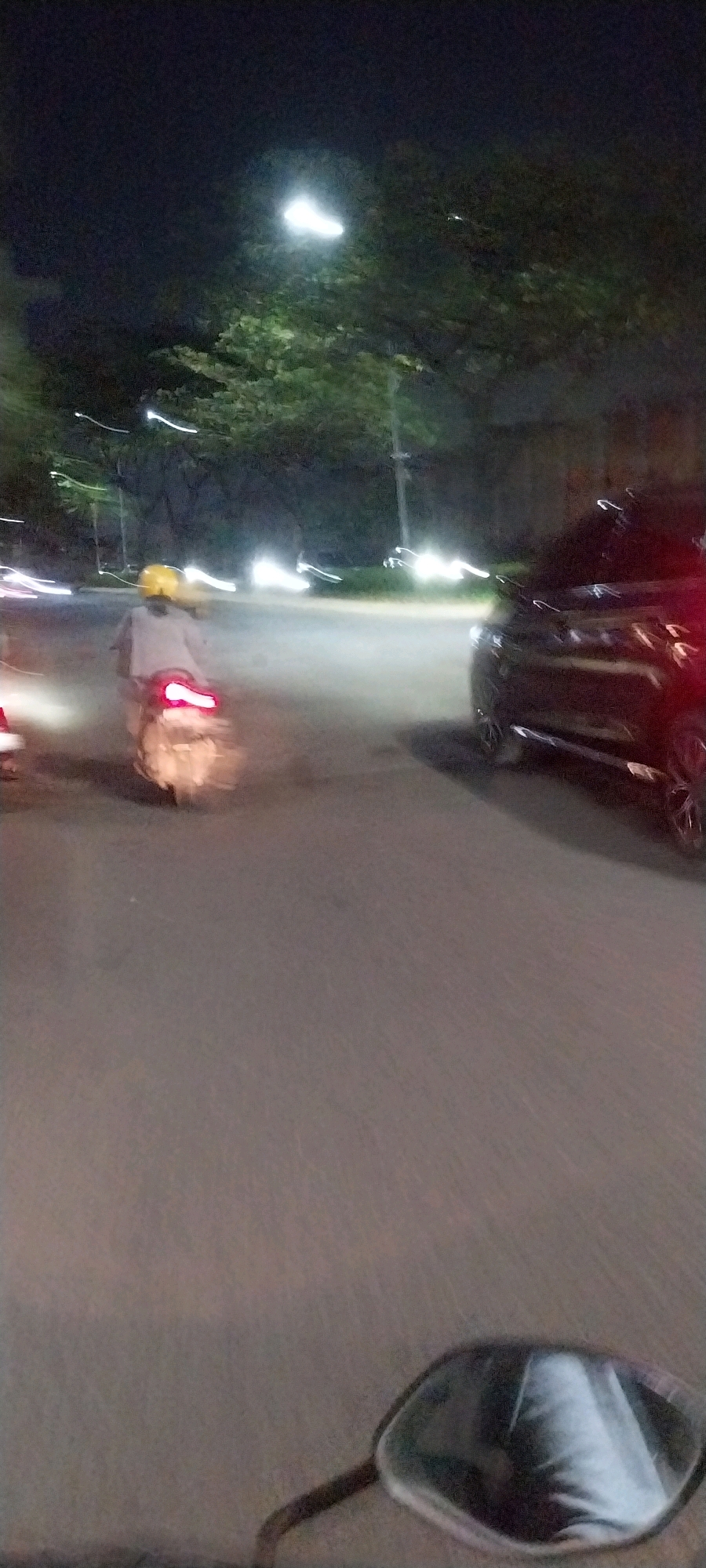 In Medang, Indonesia, at night, a person wearing a yellow helmet rides a scooter on a road. The scooter's taillight is illuminated, suggesting it is in motion. A dark-colored car is visible on the right side of the road, also appearing to be in motion due to motion blur. Streetlights and vehicle headlights cast a glow on the surroundings, illuminating trees and the road. The overall scene depicts a nocturnal street view with active traffic, characterized by motion blur due to the low-light conditions and movement.