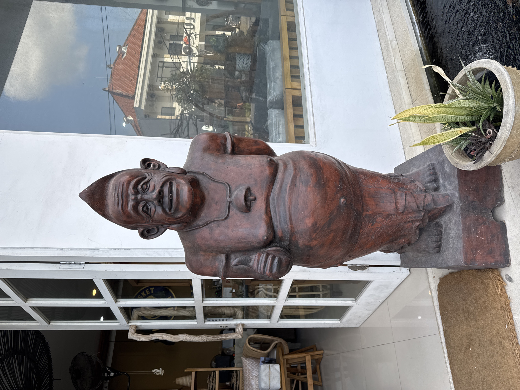 In North Kuta, Indonesia, a reddish-brown wooden statue of a man stands outside a building with a white facade and a glass door. The statue is life-sized and depicts a portly figure with a pointed hairstyle, large ears, and a smiling expression, giving a thumbs-up with its right hand. A bell hangs from a cord around its neck. The statue rests on a dark rectangular base. To the left of the statue, a brown coir doormat is placed on the tiled floor, and an open white door reveals shelves filled with items inside the building. To the right of the statue, a potted plant with long, green and yellow leaves sits next to a dark water feature. The glass door of the building reflects the sky and a building with a terracotta roof.