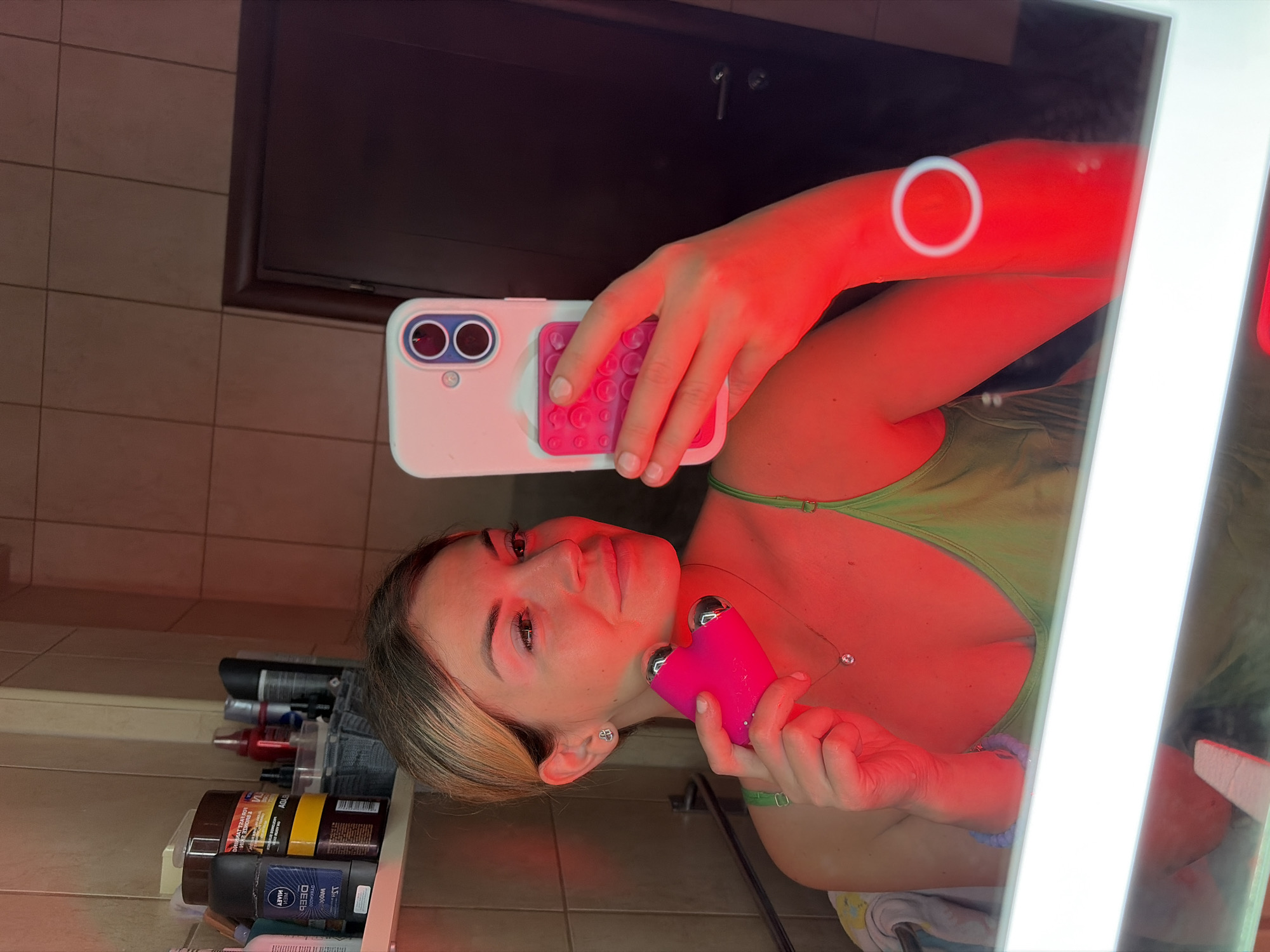A woman with blonde hair tied back is holding a pink heart-shaped device and a white smartphone in a bathroom. She is taking a selfie in a mirror, and the room is illuminated by red light. She is wearing a green tank top and a delicate necklace. The bathroom shelves are visible in the background, stocked with various toiletries and cosmetics. The scene appears to be an indoor domestic setting, likely a personal bathroom.
