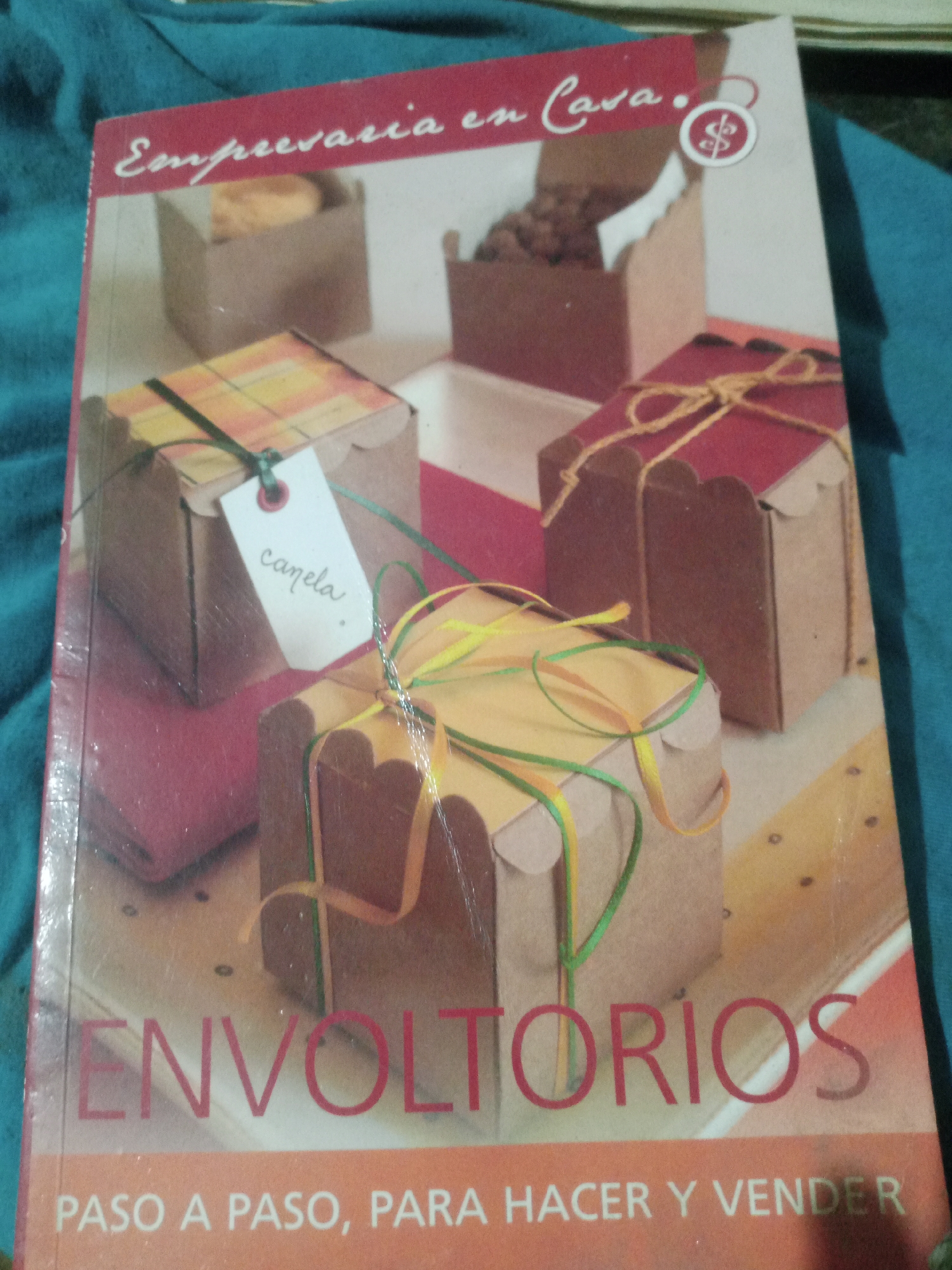 The image shows the cover of a book titled "Empresaria en Casa" and "Envoltórios Paso a Paso, Para Hacer y Vender". The cover features an illustration of various wrapped gift boxes, some decorated with ribbons and tags. There is a small circular graphic with a dollar sign on the upper right side of the cover. The book is resting on a turquoise fabric surface. The image does not provide any information about the city of Santa Ana, Venezuela, or any people present.