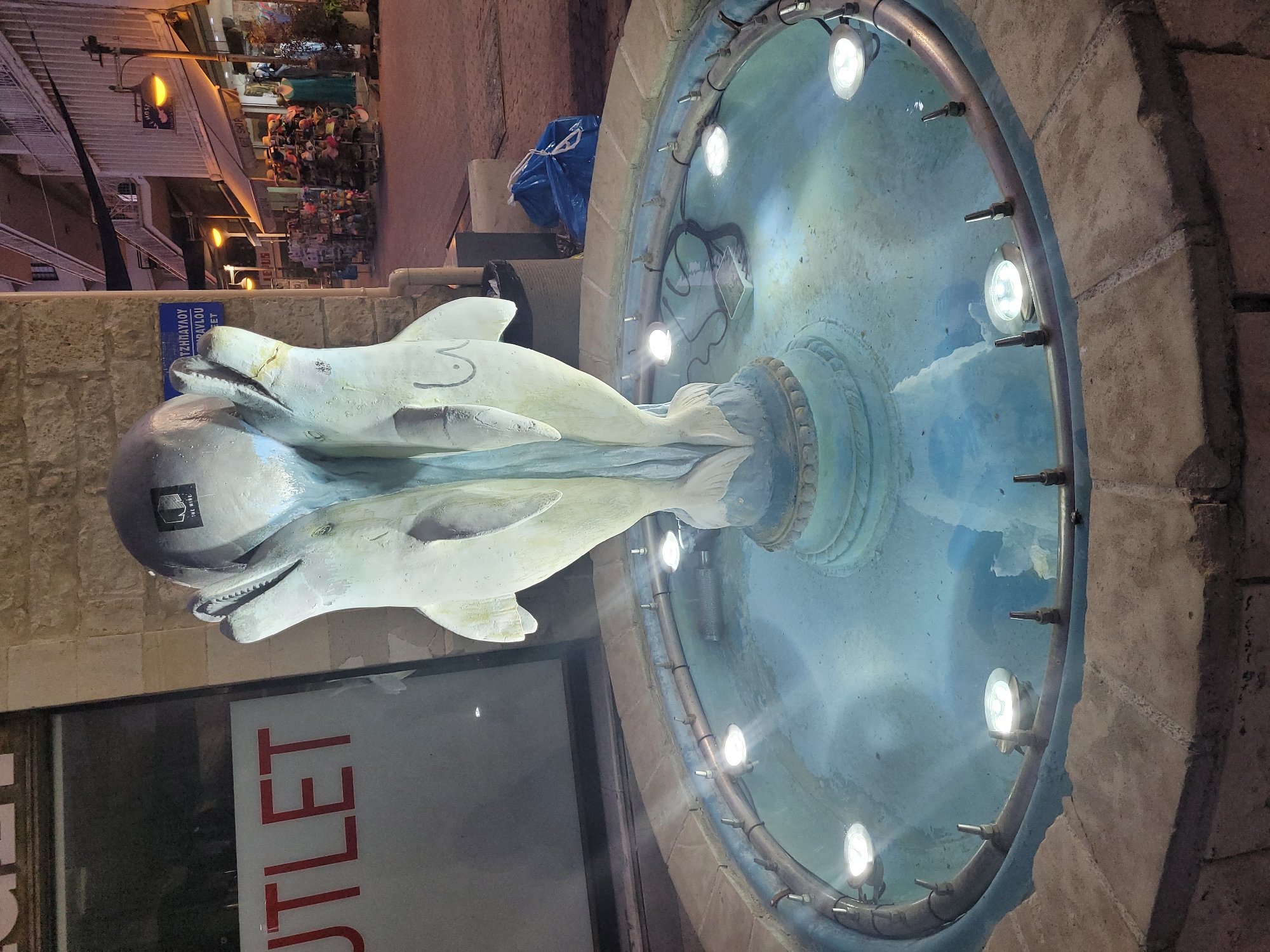 The image shows a fountain with a sculpture of two dolphins in the city of Limassol, Cyprus. The sculpture is made of a light-colored material and is positioned in the center of the fountain. The fountain is filled with blue water and illuminated by several underwater lights. In the background, there is a building with windows and a street with shops. The scene appears to be at night, as indicated by the artificial lighting. There is a sign on the building that says "OUTLET". A blue garbage bag is visible on the ground next to the fountain.