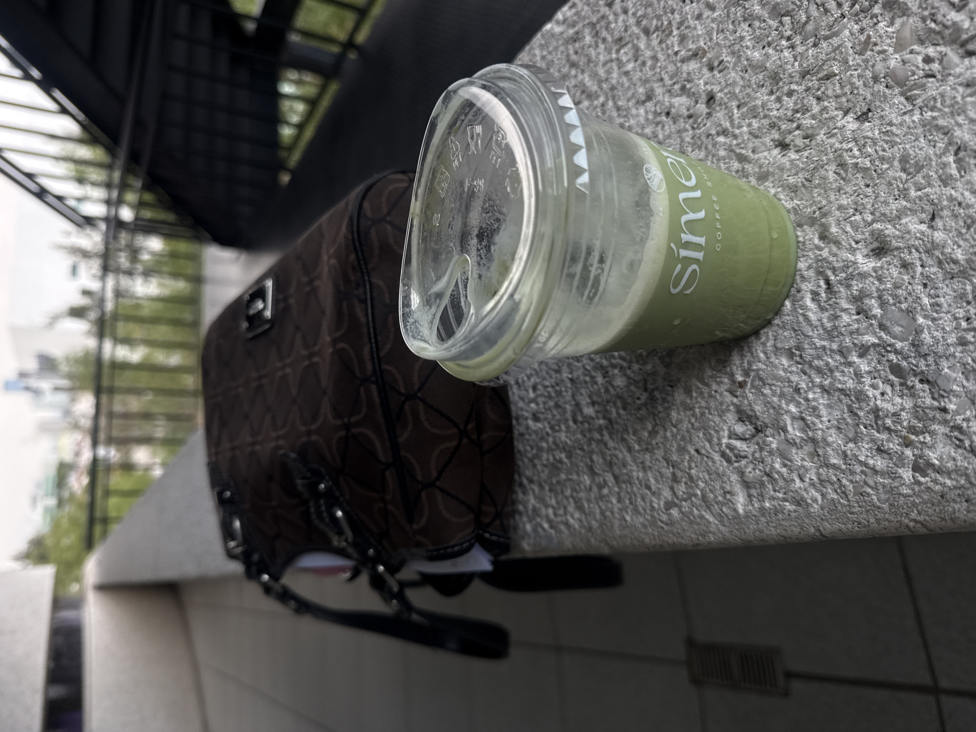A brown purse with a diamond pattern and black handle is positioned on a concrete ledge. Next to the purse is a clear plastic cup with a green liquid. The cup is labeled "Sime" and "Coffee Shot". The ledge appears to be part of a building exterior, with a blurred cityscape visible in the background. There are vents underneath the ledge.
