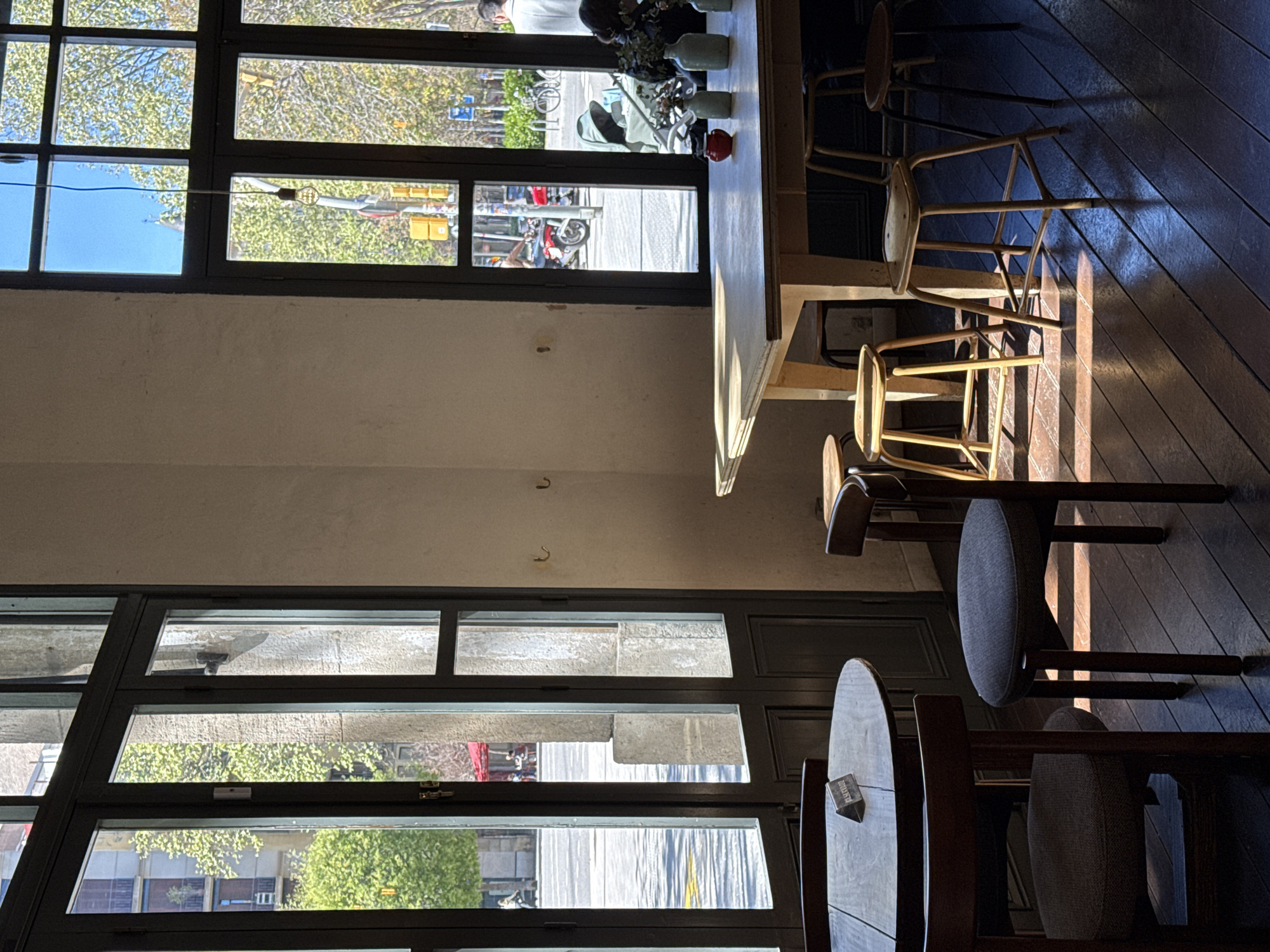 The image shows the interior of a restaurant or café in Barcelona, Spain. A large window with multiple panes provides a view of the street outside, where pedestrians, vehicles, and buildings are visible. The interior features a long counter or bar with stools, and additional tables and chairs are arranged on a wooden floor. Sunlight streams through the window, creating strong shadows and highlighting the details of the furniture. The overall ambiance suggests a casual and modern establishment.