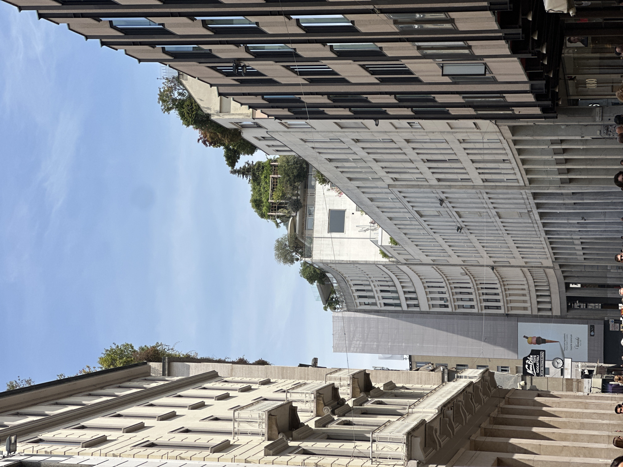 The image captures a street scene in Milan, Italy, featuring a mix of architectural styles. On the left, a pale stone building with ornate balconies lines the street. To its right, a modern, cream-colored curved building dominates the mid-ground, adorned with numerous windows and some greenery on its rooftop. Further to the right, a darker, multi-story building with vertical dark brown elements and windows stands prominently. The sky is a clear, pale blue. In the mid-ground, a large billboard displays ice cream and text in Italian. The lower portion of the image shows a section of the sidewalk with several people walking.