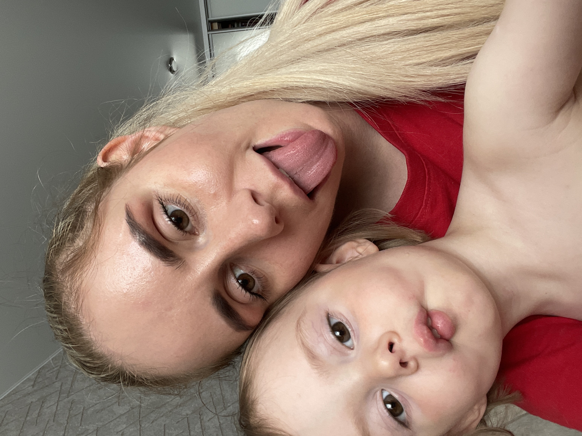 A selfie shows a blonde-haired woman and a young child. The woman, positioned at the top, sticks out her tongue towards the camera. The child, located near the bottom, also has a slightly protruding tongue. Both are wearing red shirts. The background appears to be an interior setting with a gray wall. There is no evidence to indicate this image was taken in Vladivostok, Russia.