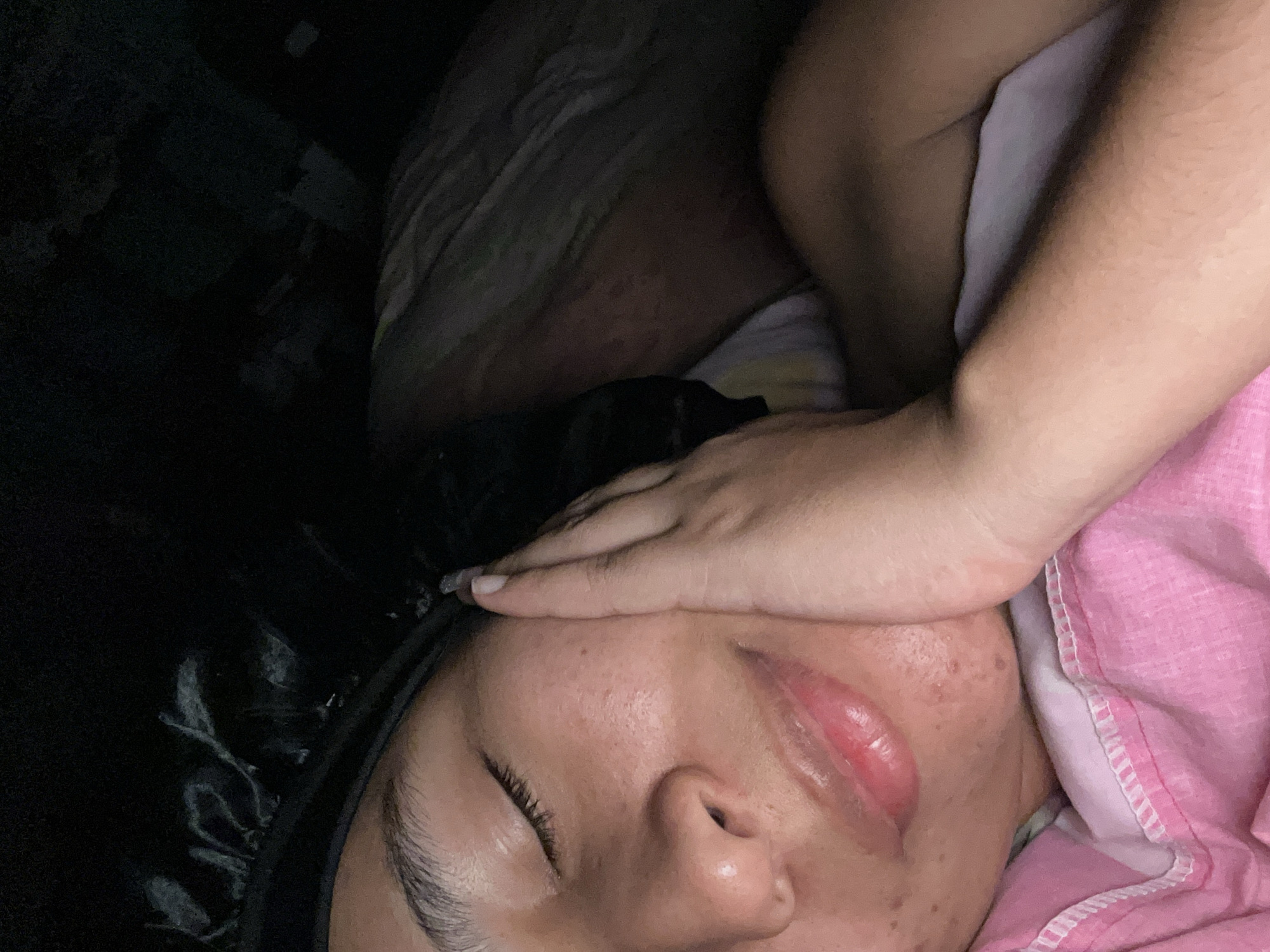 A close-up, tilted-angle photograph captures a sleeping person, likely female, covered in bedding. Her skin is visible with details like pores and minor blemishes. She wears a black head covering, possibly a bonnet. Her face is partially obscured by a hand placed across her forehead. The bedding includes a pink covering and patterned sheets. The left side of the image is dominated by dark, undefined shapes. Juan Griego, Venezuela, is not discernible from the image.