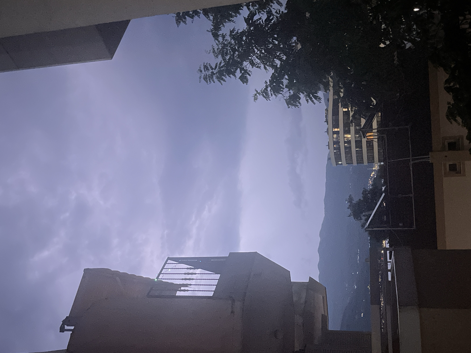 The image depicts a nighttime or twilight scene with a dramatic, cloudy sky. In the foreground, to the left, is a portion of a building's balcony with a black metal railing. To the right, a dark silhouette of a leafy tree is prominent. In the middle ground, a distinctive curved building with lit windows stands out against the dusky sky. Beyond this, a mountainous landscape with scattered lights, suggesting a city or town, is visible. The overall atmosphere is somber and somewhat dramatic, likely influenced by the cloudy sky and low light conditions. The scene is consistent with a view from an elevated position in Budva, Montenegro, overlooking the city and its surrounding natural landscape as evening descends.