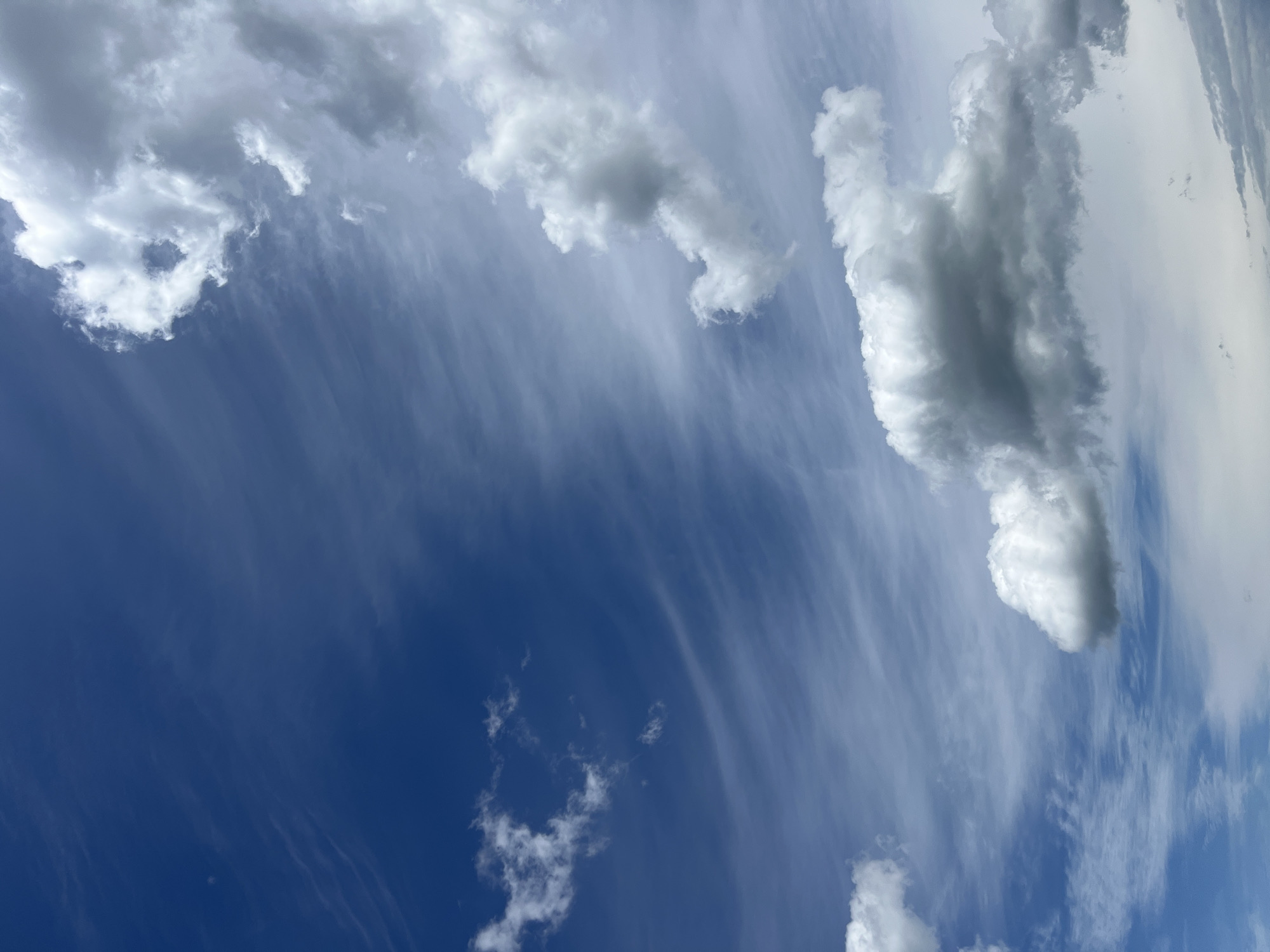 This is a vertical, close-up shot of a partly cloudy sky over Borychivka, Ukraine. The sky transitions from a deep, rich blue on the left to a lighter, hazy blue on the right. Streaky, wispy clouds with a feathery appearance are present throughout the frame, particularly in the upper left and diagonally across the center. Puffy, cumulus clouds, some with darker gray undersides indicating depth or potential for rain, are scattered across the sky. The lighting suggests it is daytime, with sunlight illuminating the white portions of the clouds and creating contrast. There are no people, structures, or ground-level objects visible. The scene is solely focused on the atmospheric conditions above Borychivka.