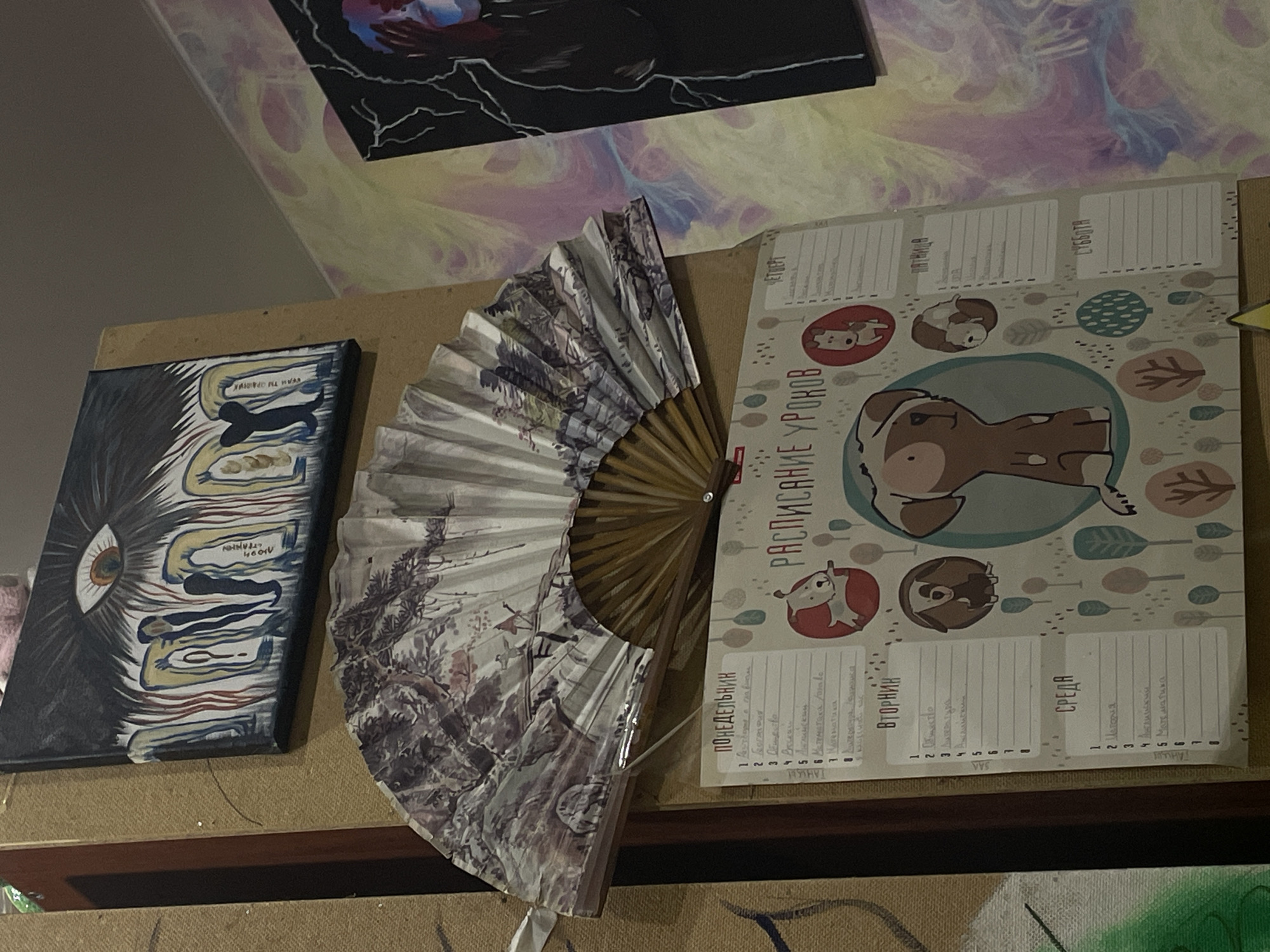 The image shows a close-up view of a wall decorated with artwork and a decorative fan. In the center of the image is a folded fan with a landscape pattern. Below the fan is a printed schedule titled "Расписание Уроков" (Schedule of Lessons), featuring cartoon dogs and trees. The schedule is organized by days of the week, with sections for Monday, Tuesday, Wednesday, and Saturday visible. To the left of the schedule, there is a painting with an eye motif and figures. To the right, another painting with abstract swirling colors and a dark silhouette of a person is visible. The overall scene suggests an indoor setting, likely a room, adorned with personal artistic expressions. The location "ЖК «Борисоглебское», Russia" is noted, which is a residential complex.