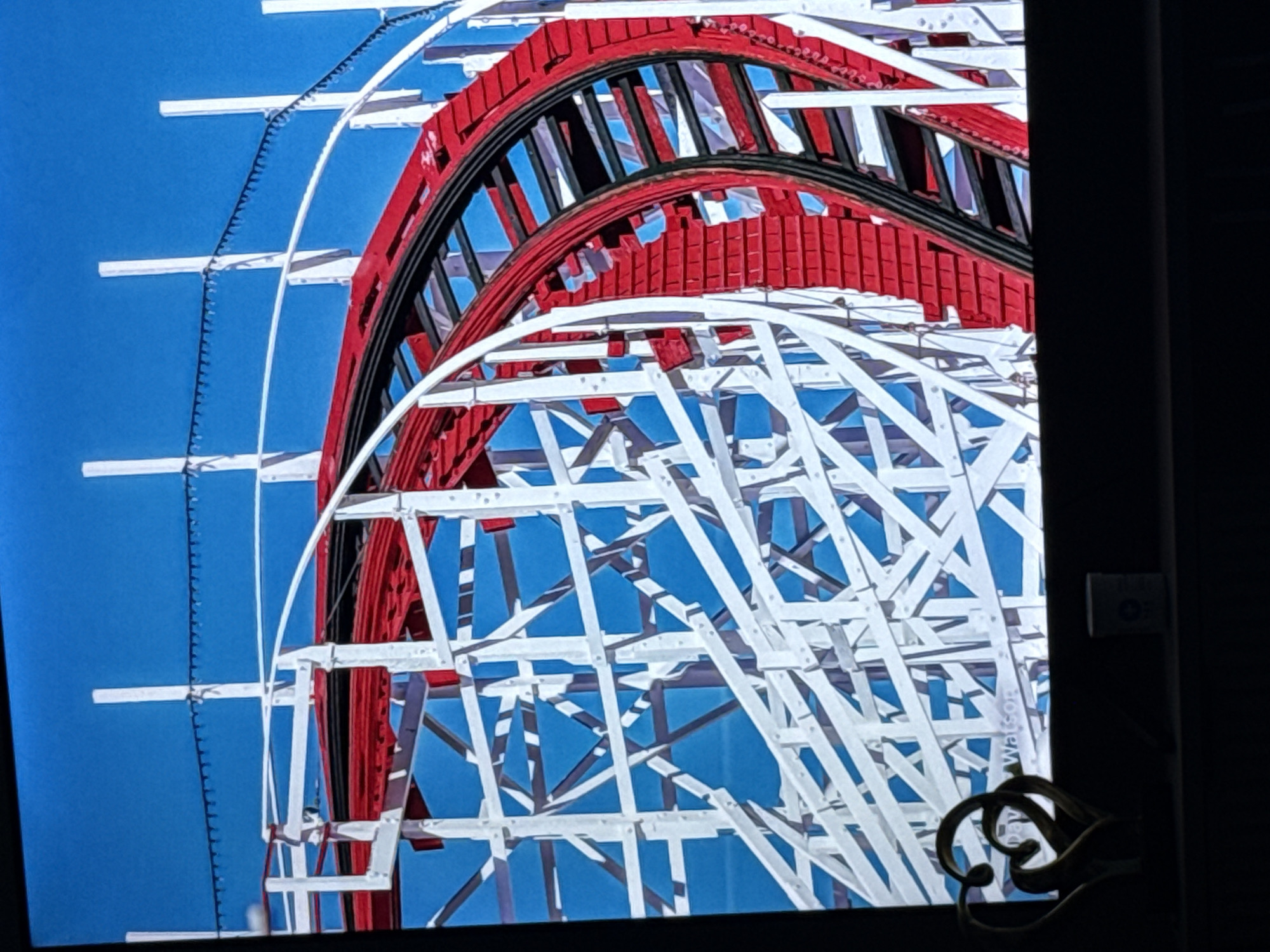 The image depicts a close-up view of a portion of the Cyclone Racer roller coaster in Long Beach, California. The coaster is characterized by its red and white painted wooden structure. The sky is a clear blue. The image is taken from an oblique angle.