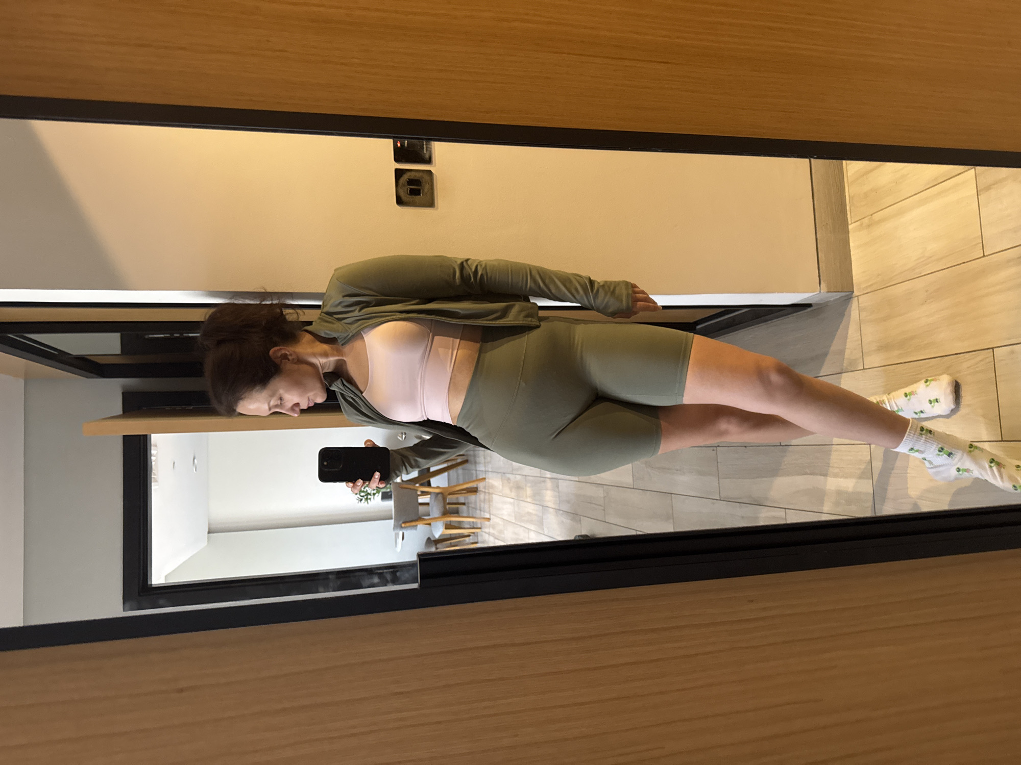 A woman is reflected in a mirror, holding a black smartphone. She wears a light pink sports bra, a green jacket, and green shorts. Her socks are white with green designs. The reflection shows a view into a room with white walls, chairs, and potted plants. The surrounding area shows a wooden wall, flooring tiles, and electrical outlets. No information implies that the location is Dubai.