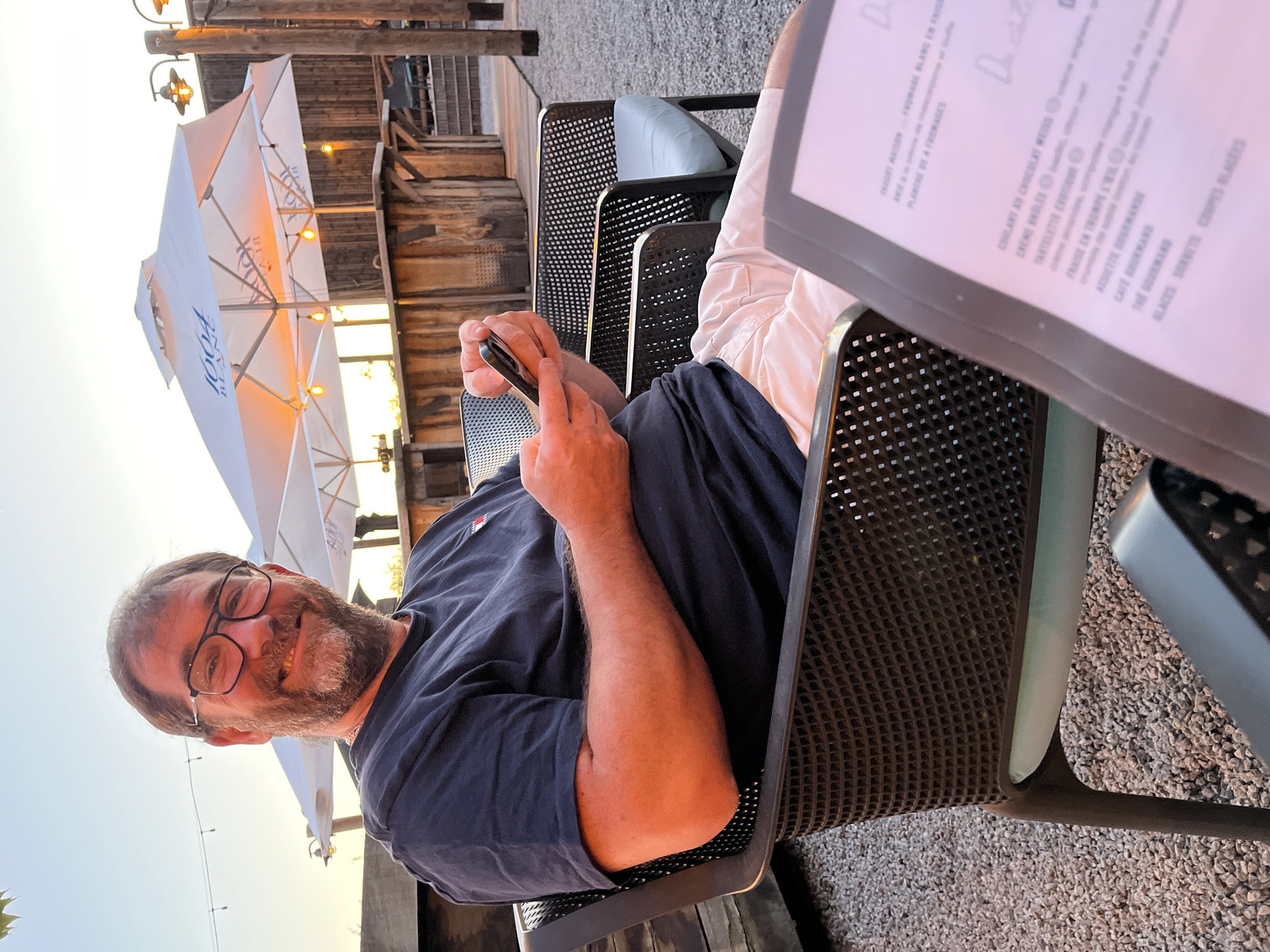 A medium shot depicts a middle-aged man seated in a metal chair at an outdoor restaurant. The restaurant is located in Andrézieux-Bouthéon, France. He is wearing glasses, a dark blue t-shirt, and white shorts, and he is holding a mobile phone. A menu is visible on the table next to him. Above, there is a large white parasol with the logo of 1664 Blanc, and behind it, a wooden building. The scene is sunlit.
