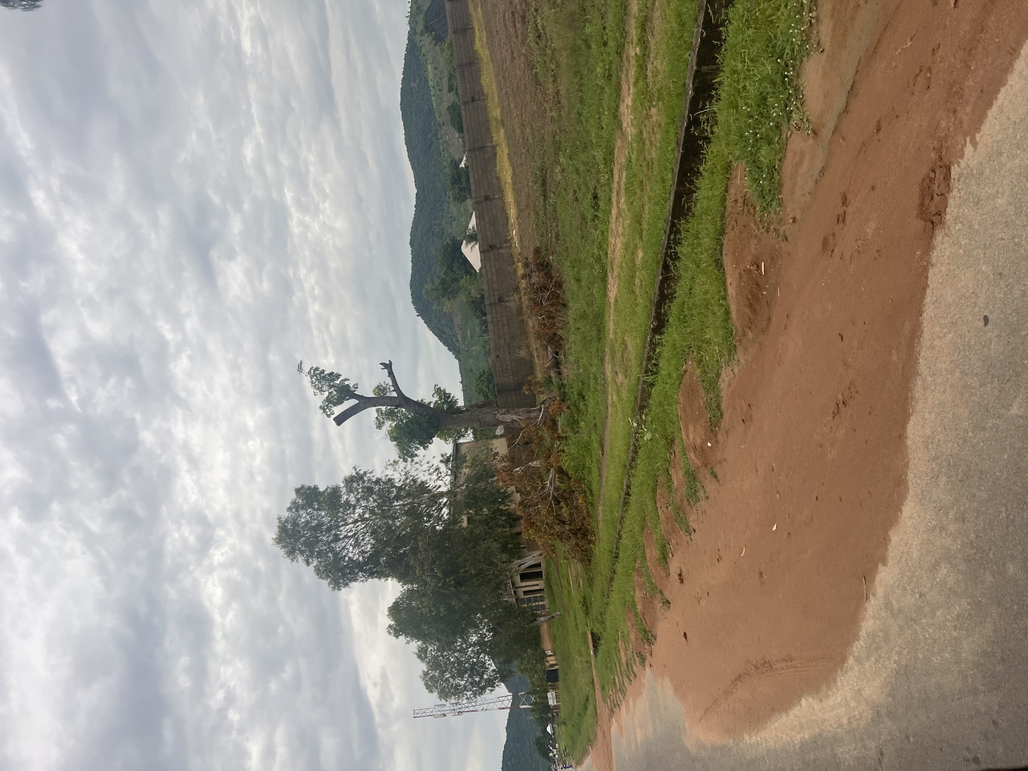 The image depicts a roadside scene in Kona Garu, Nigeria, under a cloudy sky. A paved road with a dirt shoulder runs along the left side of the frame. On the right, a grassy area is bordered by a concrete drainage channel. Beyond the grass, a low brick wall encloses a dirt field. In the background, several buildings and trees are visible, with a cellular tower standing tall to the left. Further in the distance, rolling green hills form the horizon. Scattered fallen branches lie on the ground in front of the buildings. The overall impression is one of an unkempt, rural or semi-urban environment.