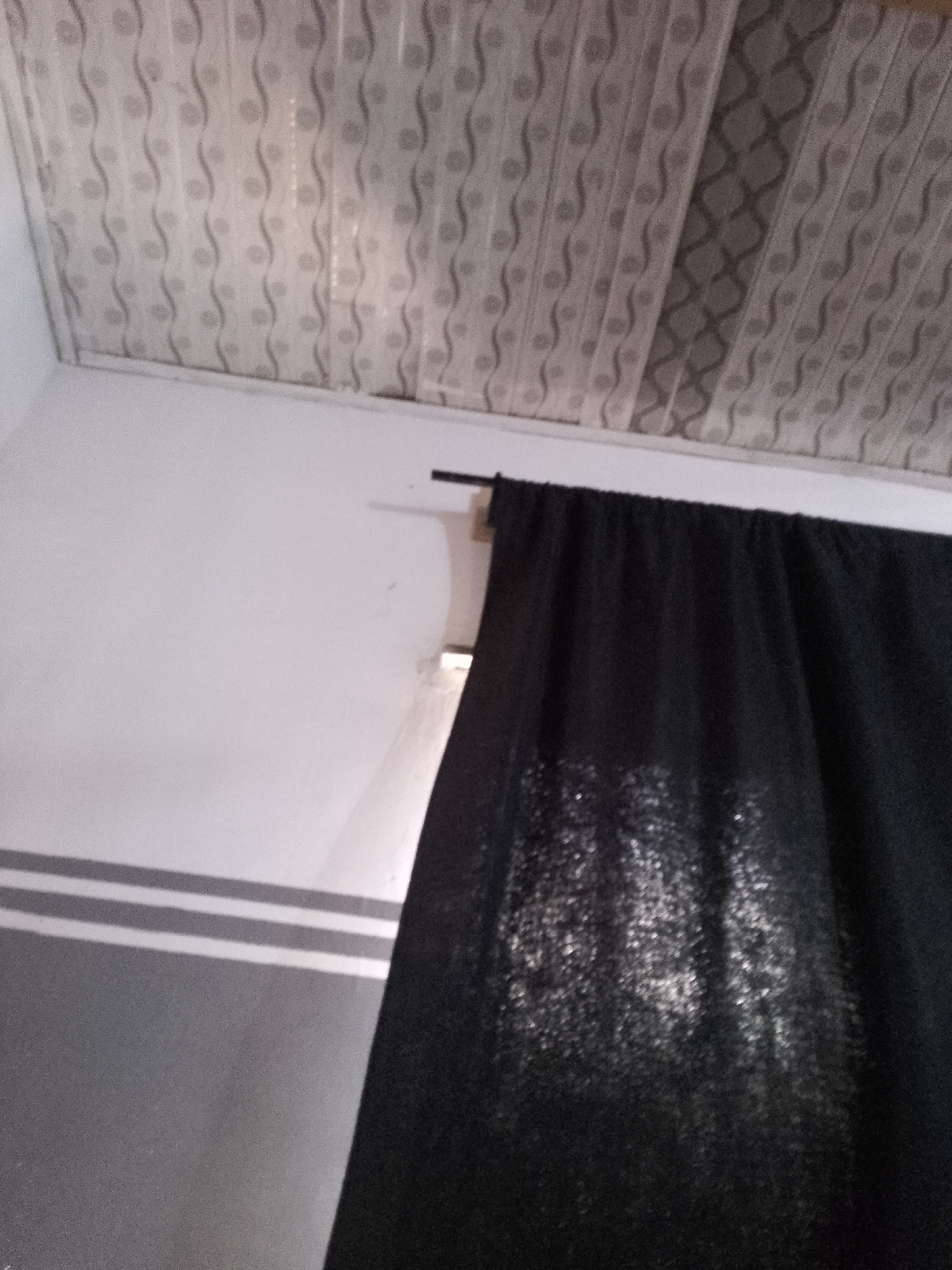 The image shows the interior of a room, likely a bedroom, in a location that cannot be determined. The ceiling is paneled with a repeating wavy pattern. A black curtain hangs from a rod, obscuring a window. The wall is white, with a section of gray wainscoting below a horizontal stripe pattern. The lighting is dim, with some light reflecting off the curtain. No people or specific actions are visible.