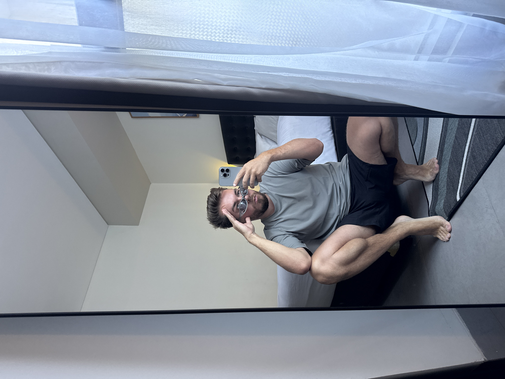 A fair-skinned man is seen in a mirror reflection. He is wearing glasses, a grey T-shirt, and black shorts. He is crouching, looking at his phone, and has one hand on his head. The background includes a white wall, a bed with white pillows, a grey and black rug, and a partially visible window with white curtains. The setting appears to be a room in North Kuta, Indonesia.