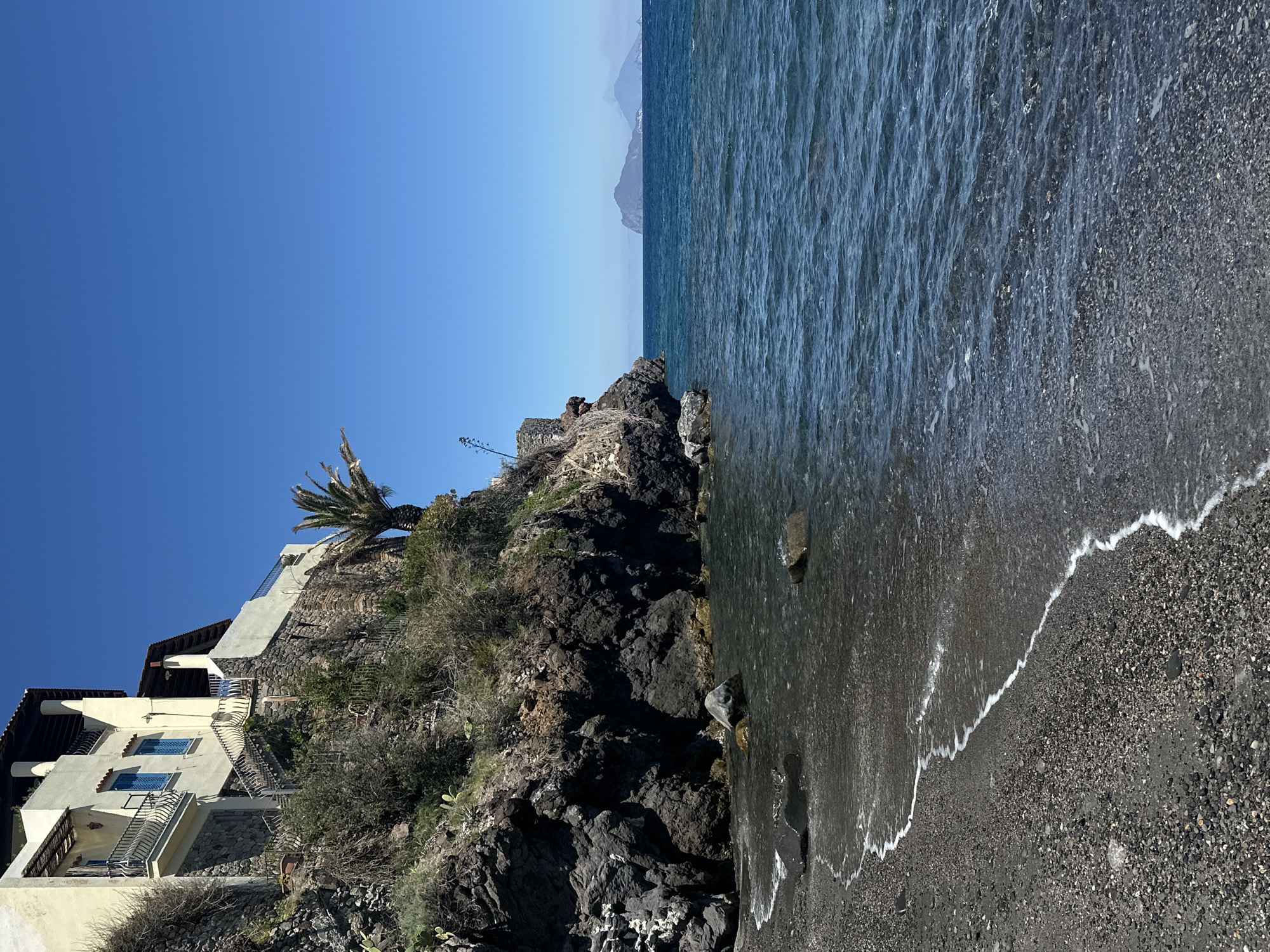 The image captures a view of the coastline in Lipari, Italy. A multi-story building with a tiled roof is constructed on a rocky cliff with vegetation. The cliff descends towards a beach with dark-colored pebbles. The sea appears turbulent and stretches to the horizon. In the distance, a faint outline of mountains is visible under a clear blue sky.