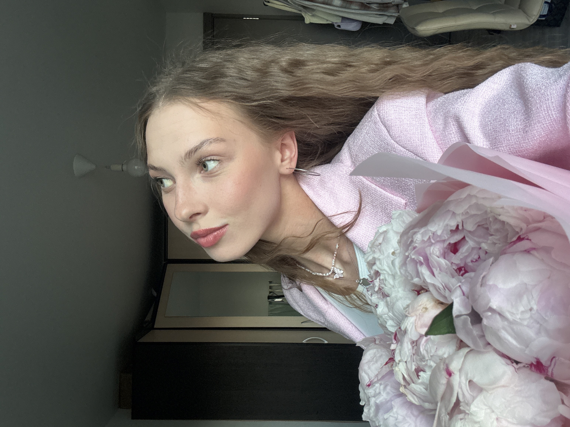 The media file is a portrait of a young woman with long, wavy blonde hair. She is wearing a light pink textured jacket over a white top and a delicate silver necklace with a flower pendant. She is holding a bouquet of large pink and white peonies, partially obscuring her chest. The woman is looking off to the left, her expression is serene. Her makeup includes subtle eyeliner and rosy blush and lipstick.
The setting appears to be indoors, possibly a home or a studio. Behind her, a dark wooden cabinet is visible, with framed pictures or artwork displayed on top. A light fixture with a bare bulb is mounted on a grey wall to her left. The overall lighting is soft, suggesting it might be daytime, possibly late afternoon or evening, given the muted light. The background is slightly blurred, drawing attention to the subject and the flowers. The image quality is good, with clear details of the woman's features and the texture of her clothing and the flowers. No text is visible in the image. The woman's posture and the presence of the flowers suggest a moment of quiet reflection or anticipation. The location context of Perm, Russia is provided, but there are no specific visual cues within the image to confirm this location.