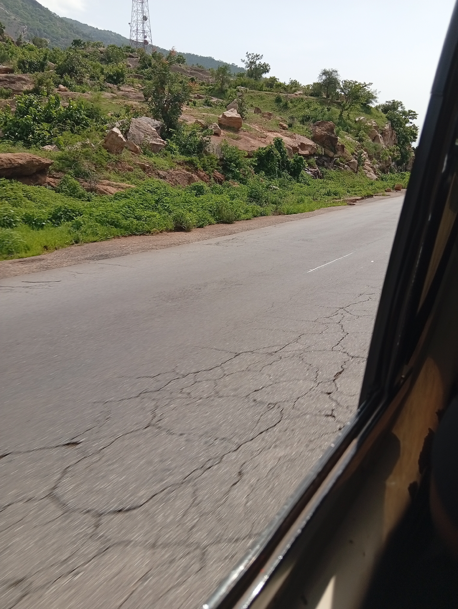 This media file captures a roadside view from inside a vehicle, likely taken in Rimin Ziem, Nigeria. The scene depicts a paved road with visible cracks, suggesting it's well-traveled. To the left of the road, a lush green hillside rises, dotted with various types of vegetation, including bushes and trees. Large rocks and boulders are interspersed on the hillside, adding texture to the landscape. In the background, a tall telecommunications tower is visible on the crest of the hill, indicating a developed area or a strategic location. The sky is clear and bright, suggesting a daytime scene with fair weather. The perspective from inside the vehicle, with a portion of its interior frame visible on the right, implies the image was taken while in motion, perhaps on a journey. The overall mood is serene and observational, focusing on the natural landscape and the infrastructure present in the region.