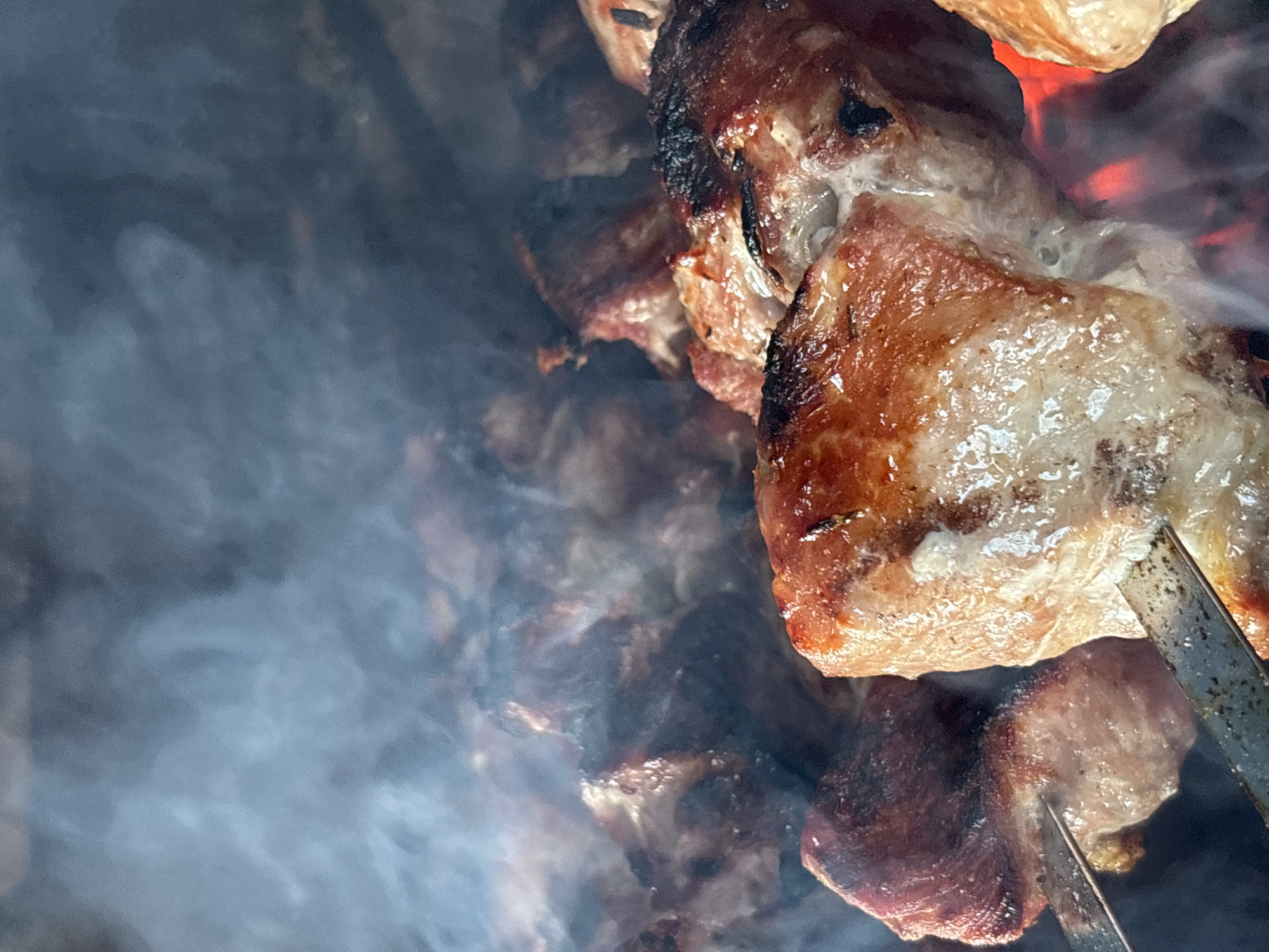 The image depicts pieces of meat skewered on metal rods, being grilled over an open fire. Smoke rises from the fire, obscuring some of the meat. Some pieces of meat appear to be fully cooked with dark char marks on the edges, while others are partially cooked. The location is unspecified, and there is no indication of the city of Chaikovsky, Russia. No people are visible in the image.