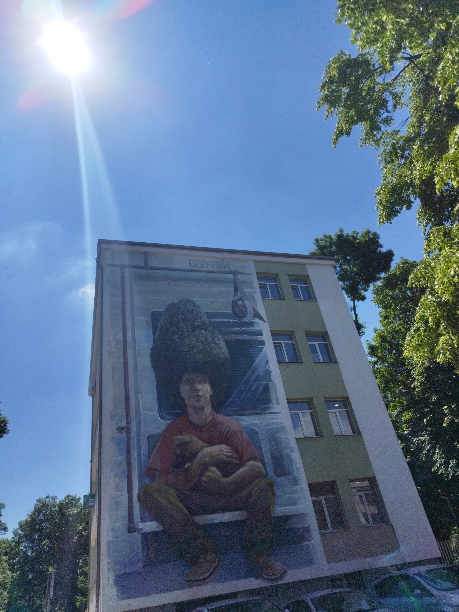 The image shows a low-angle view of a building with a large mural on its side. The mural depicts a man seated, holding a cat, with a large, bushy tree above his head as if it were his hair. The building is multi-storied with windows visible on the side where the mural isn't painted. The sky is clear and blue with the sun shining brightly. Green trees surround the building, partially obscuring the view. Several cars are parked in front of the building. This mural is located on a building in Sofia, Bulgaria.