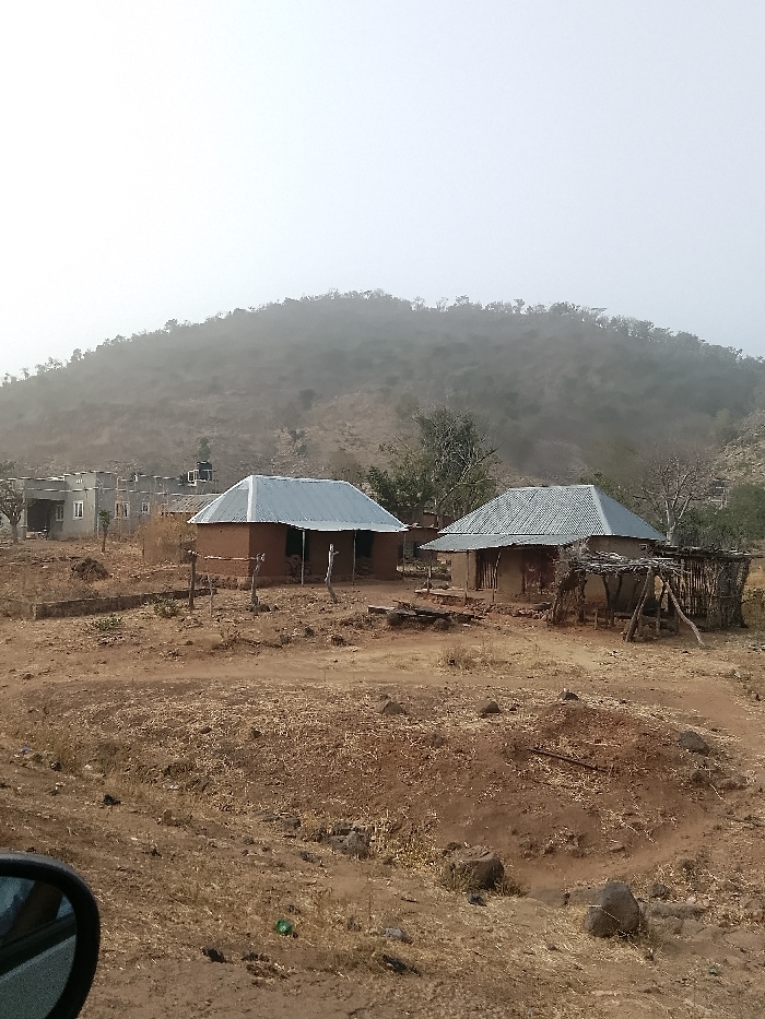 The image presents an arid landscape in Jibam, Nigeria, under a hazy, pale sky. The terrain consists predominantly of dry, reddish-brown soil with sparse, desiccated vegetation and scattered rocks. A large, tree-covered hill, obscured by atmospheric haze, rises in the background.
In the mid-ground, two traditional single-story structures with reddish-brown earthen walls and corrugated metal roofs are situated. The structure on the left features two visible rectangular openings. The structure on the right has a dilapidated, open-sided wooden annex with a partial brush roof attached to its immediate right side. A small, rudimentary wooden fence stands in front of the left structure.
Further to the left background, a collection of more contemporary multi-story buildings, likely constructed of concrete, are visible; one features a black water storage tank on its roof. The immediate foreground shows a circular depression in the earth, edged with rocks, along with more scattered dry vegetation and debris. A partial view of a vehicle's side mirror is visible in the lower left corner. No persons or animate subjects are depicted in the scene.