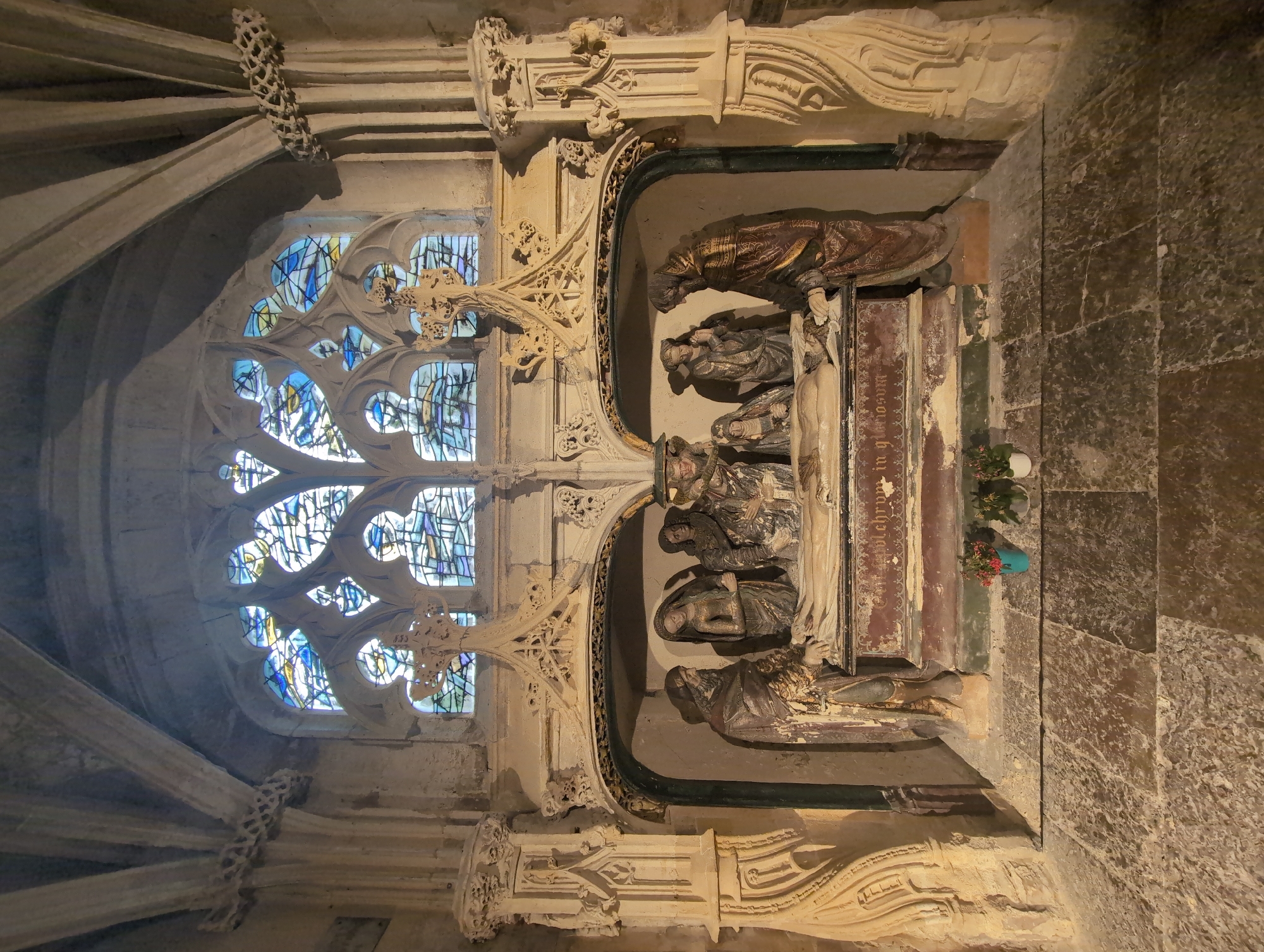 The image shows a scene inside a stone structure, likely a church or chapel, in Le Tréport, France. In the center, a sculpted effigy of a deceased person lies on a tomb. Surrounding the effigy are several figures, also sculpted, appearing to be mourners dressed in robes. The tomb is ornate, with decorative carvings and an inscription. Behind the tomb, a stained-glass window with a Gothic architectural frame is visible. The surrounding architecture is made of stone, with ribbed vaulting and decorative carvings. The floor is paved with large stone tiles. Small potted plants are placed in front of the tomb. The overall scene depicts a funerary monument.