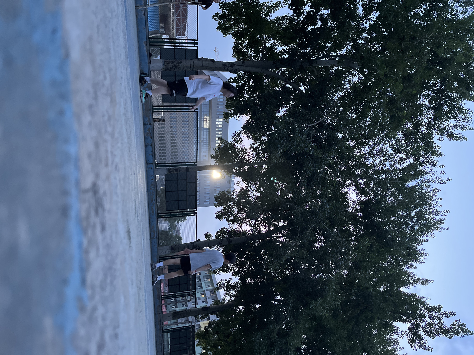 The image depicts two individuals on a basketball court, likely during an evening or twilight setting given the soft lighting and the sky's color. One person, in the foreground, is wearing a white t-shirt and black shorts, actively engaged in playing basketball. Their posture suggests movement, possibly jumping or reaching for the ball. The other individual, further in the background, is also dressed in athletic wear and appears to be participating in the game.
The basketball court has a light-colored surface with blue markings along the edges. A chain-link fence encloses the court, behind which a cityscape is visible. Tall buildings with illuminated windows suggest an urban environment, and the presence of trees with dense foliage adds a natural element to the scene. The sky is a pale blue, indicating dusk or dawn. The overall atmosphere seems relaxed and focused on the activity. No visible text is present in the image. The setting is likely an outdoor public basketball court in Daxing District, China, consistent with the provided location context.