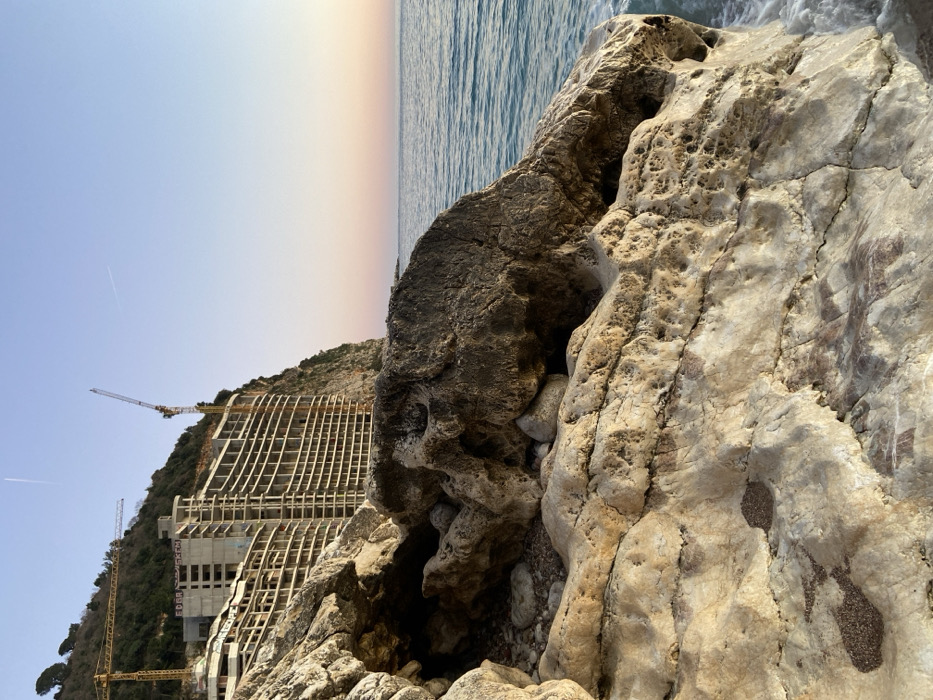 The image presents a rugged coastal landscape dominated by a rocky foreground and a large construction site on a cliff overlooking the sea. In the immediate foreground, a mass of light-colored, heavily textured rock formations with visible crevices and minor sand deposits occupies the lower half of the frame. To the right, clear blue-green ocean water laps against these rocks, exhibiting minimal wave action. A steep, vegetated cliff rises in the midground, serving as the foundation for multiple large, multi-story concrete structures in various stages of construction. These incomplete buildings feature extensive exposed frameworks and numerous horizontal tiers, likely balconies. Two prominent yellow tower cranes are positioned on the cliff alongside the buildings, indicating ongoing construction activity. The sky above is a gradient of pale blue to light orange and pink near the horizon. A single white aircraft contrail is visible in the upper left quadrant of the sky. No individuals are present. The specific location in the city of Unknown, Unknown is a rocky shoreline adjacent to significant coastal development.