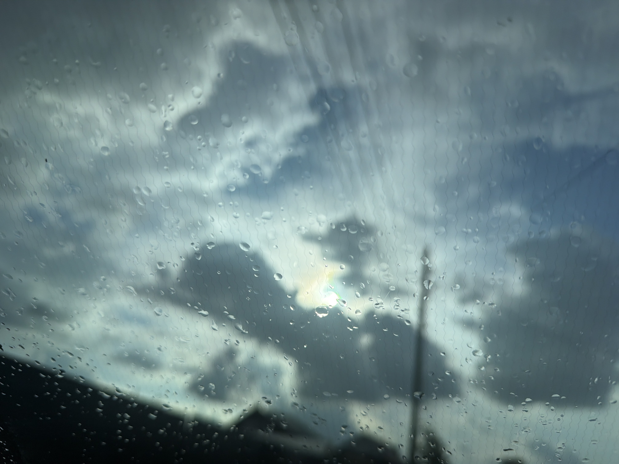The image depicts a cloudy sky seen through a rain-streaked window, possibly a car windshield, with the sun peeking through a gap in the clouds, creating a rainbow effect. In the foreground, raindrops distort the view and create a texture of ripples and streaks across the glass. The lower portion of the image shows a dark, silhouetted landscape, possibly trees or buildings, against the brighter sky. The presence of a utility pole is faintly visible on the right side of the frame. There are no people or discernible actions. The scene appears to capture a moment during or immediately after a rain shower, with the light suggesting it is daytime. The specific location in Chaikovsky, Russia, cannot be determined from the visual information alone.